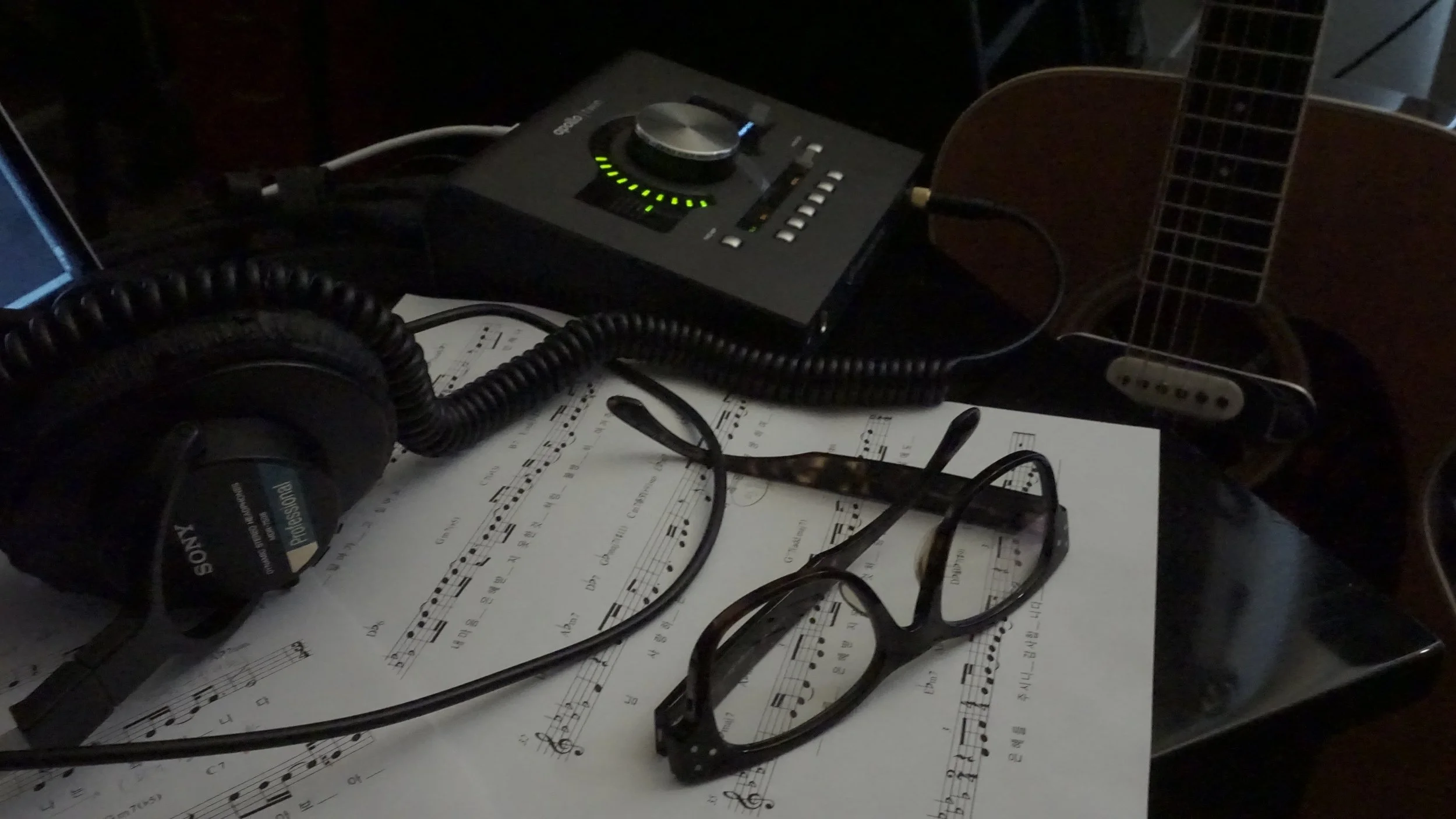 Laptop displaying a guitar chord diagram on its screen, positioned on a wooden desk with an open book nearby.