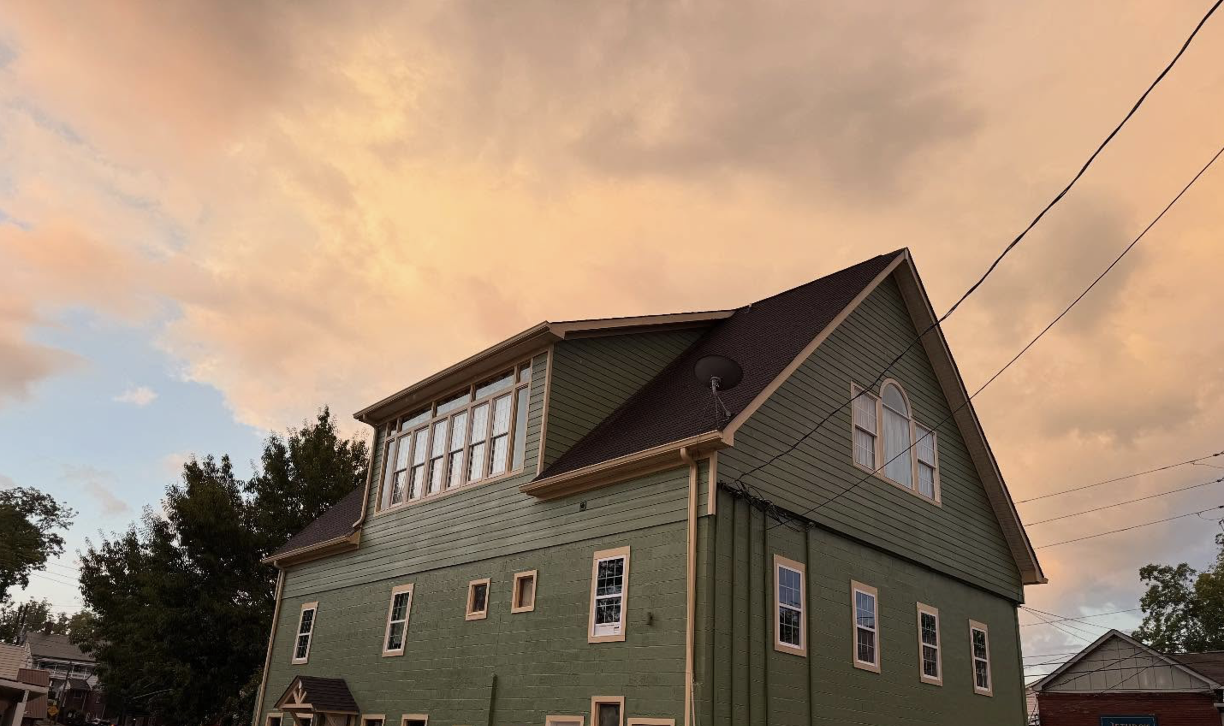 Green house with multiple windows, pointed roof, and attached electrical wires against a sunset sky with orange clouds.