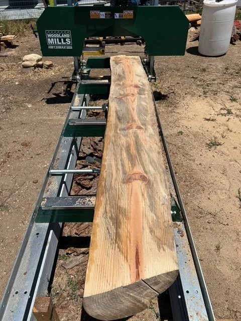 A large wooden slab on a green WoodMills thickness planer machine outdoors, with dirt ground, rocks, and a white barrel in the background.
