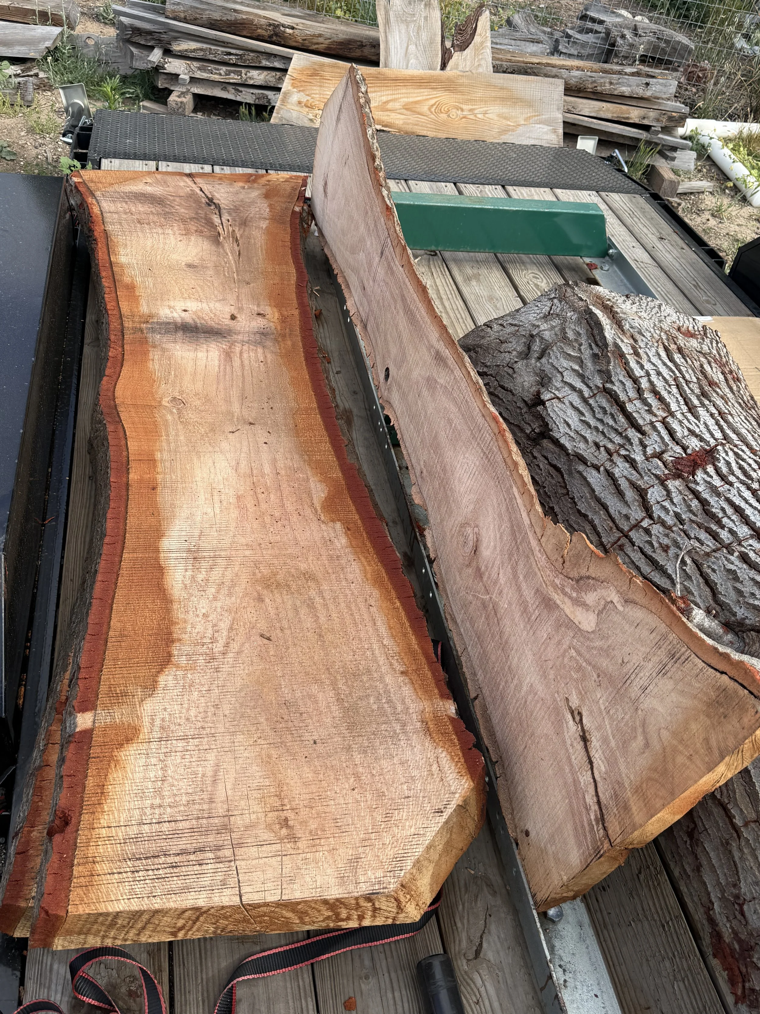 Several large cut logs of wood resting on a wooden platform outdoors, with some bark on the logs and others cut smooth. There are additional logs and wood pieces in the background.