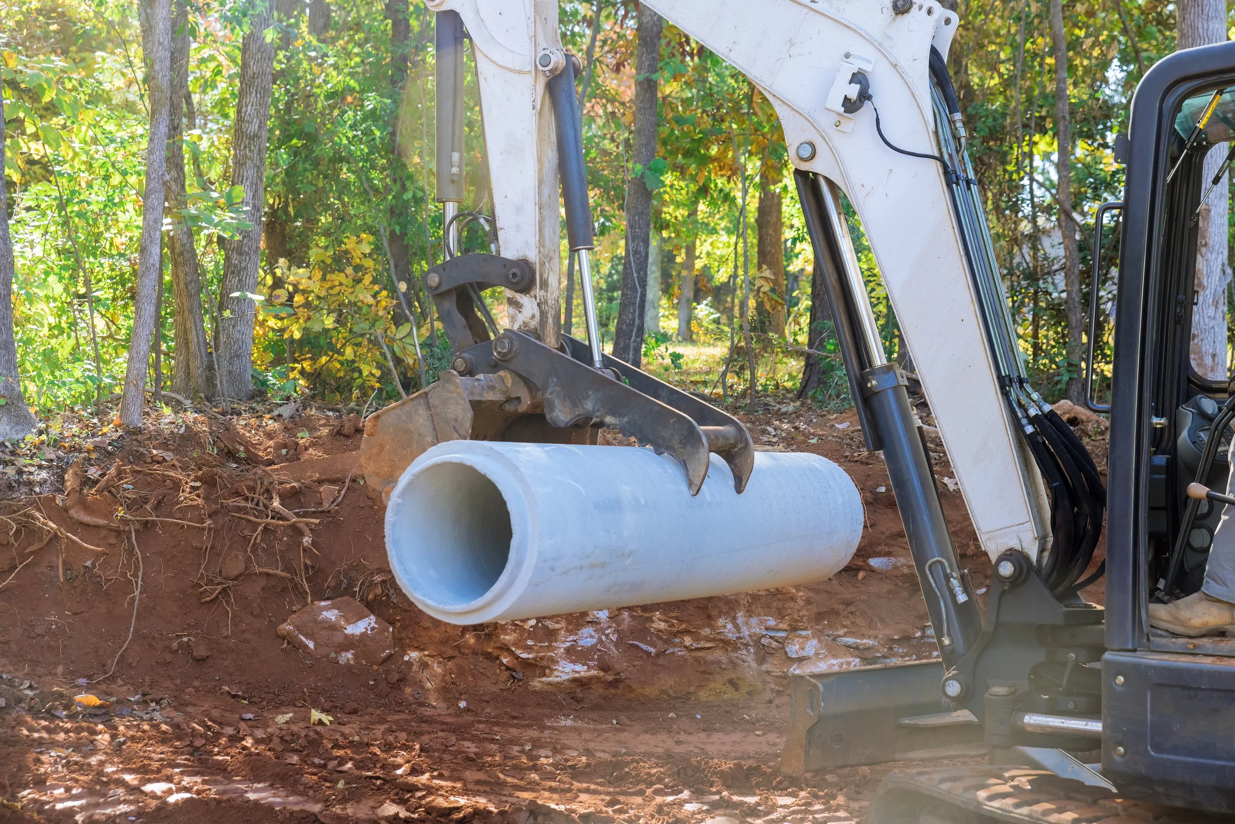 Digger placing a large gray pipe into a dug-out area in a wooded construction site.