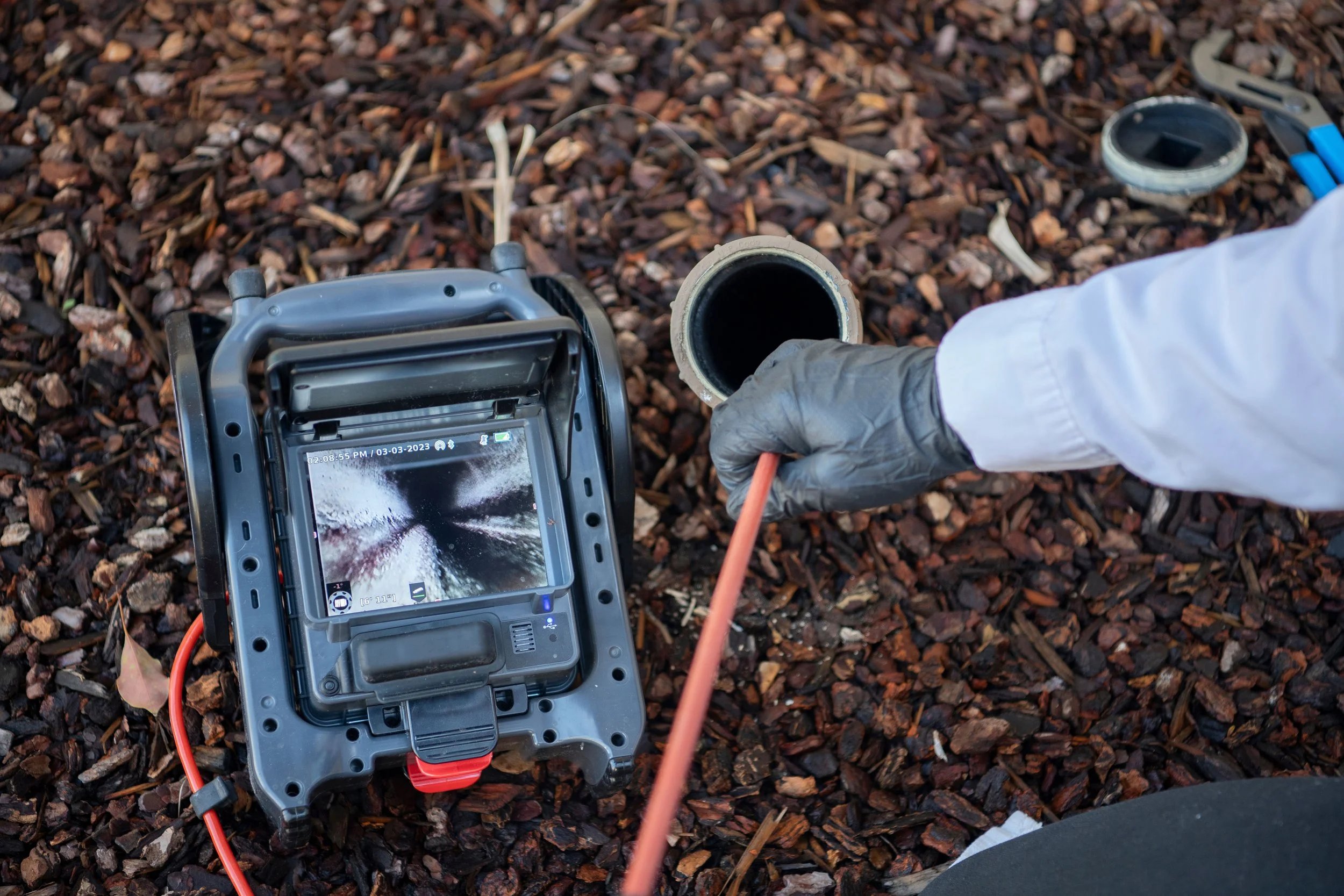 Worker in white coat and gray gloves is using a pipe inspection camera on a pipe underground, with the camera display showing inside the pipe, on a gravel surface.