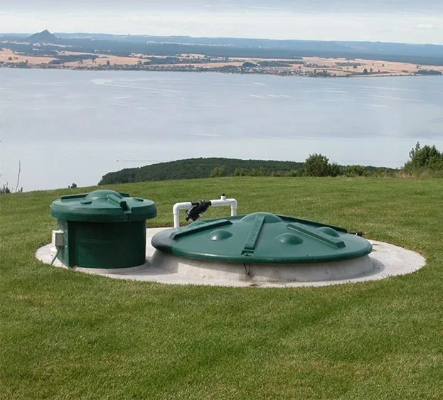 Ground-level view of a septic tank with green lids and a visible pipe, located on a grassy hill with a large body of water and distant landscape in the background.