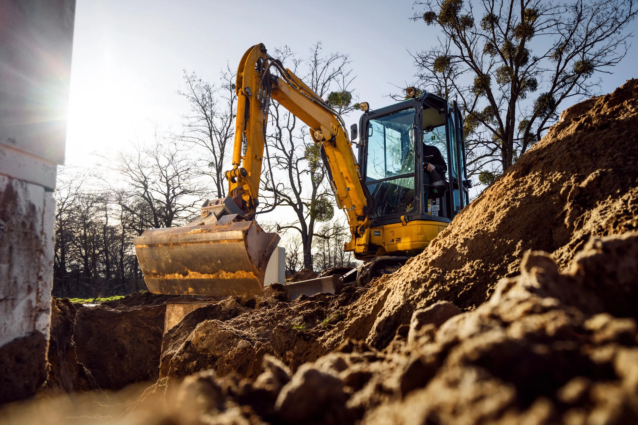 A yellow excavator digging into the soil at a construction site during daylight, with bare trees and a partly cloudy sky in the background.