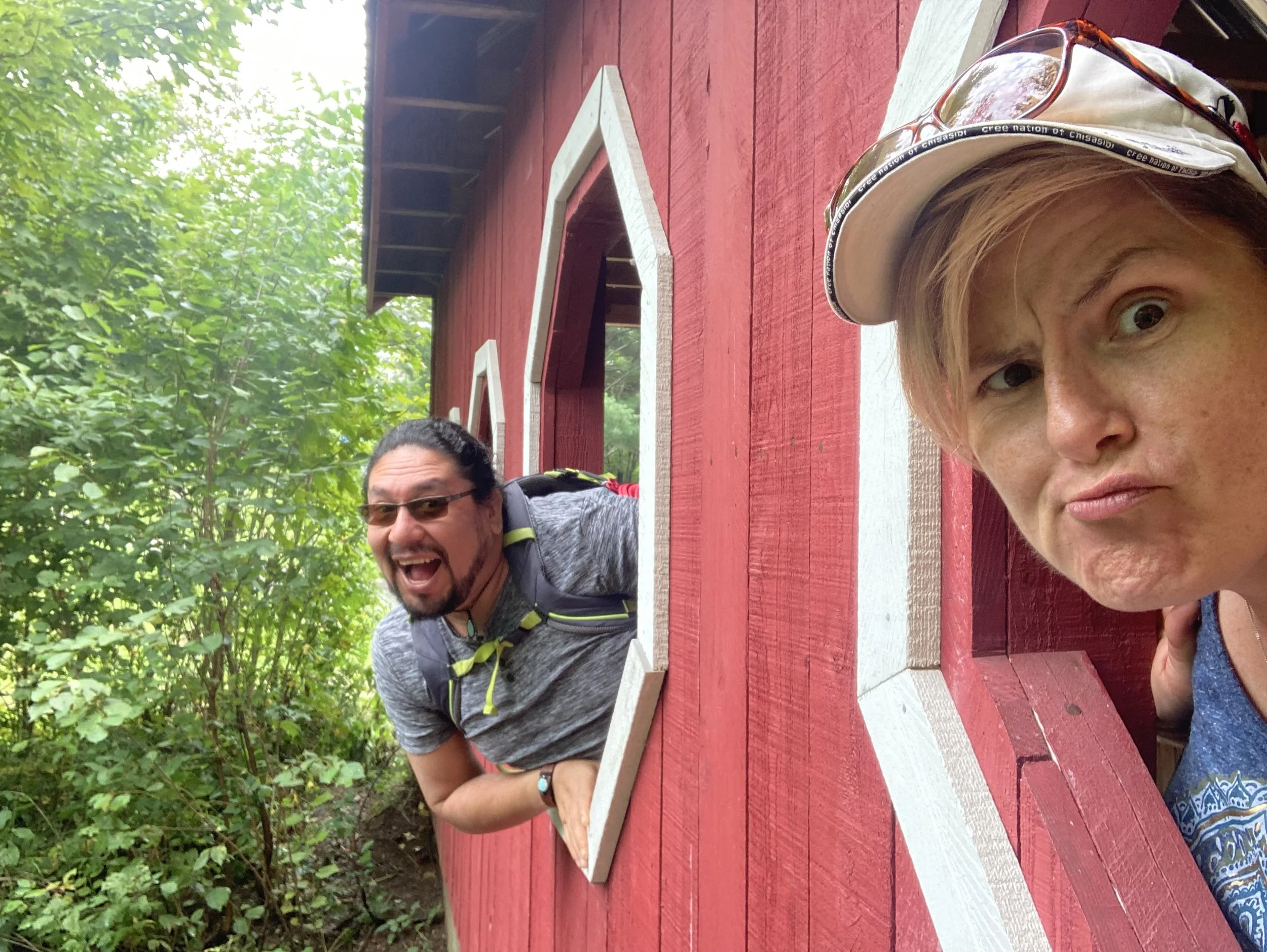 A woman and a man leaning out of a  wooden window in a red structure, surrounded by greenery, making funny faces at the camera.