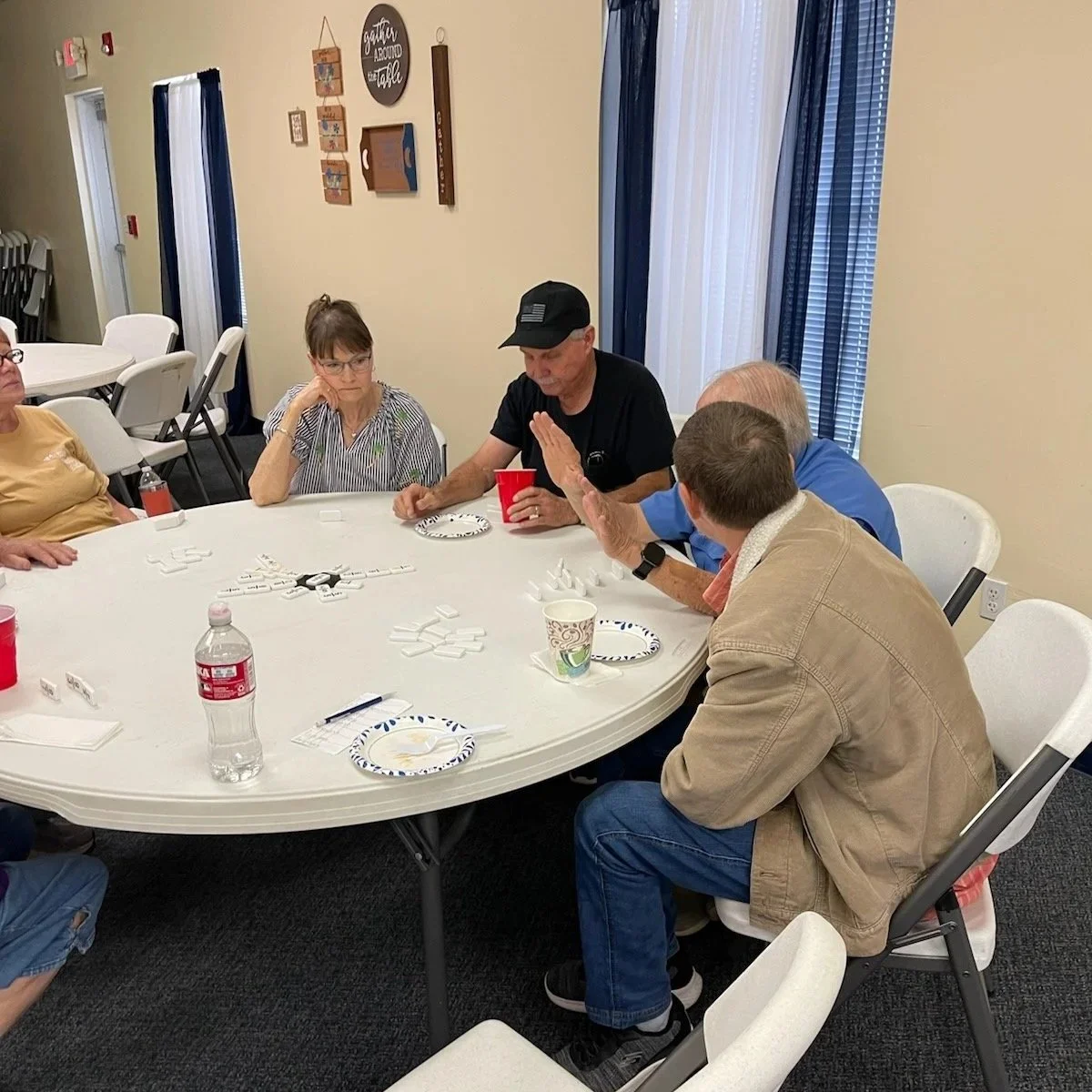 Group of people sitting around a table playing dominoes in a room decorated with signs and hanging art.