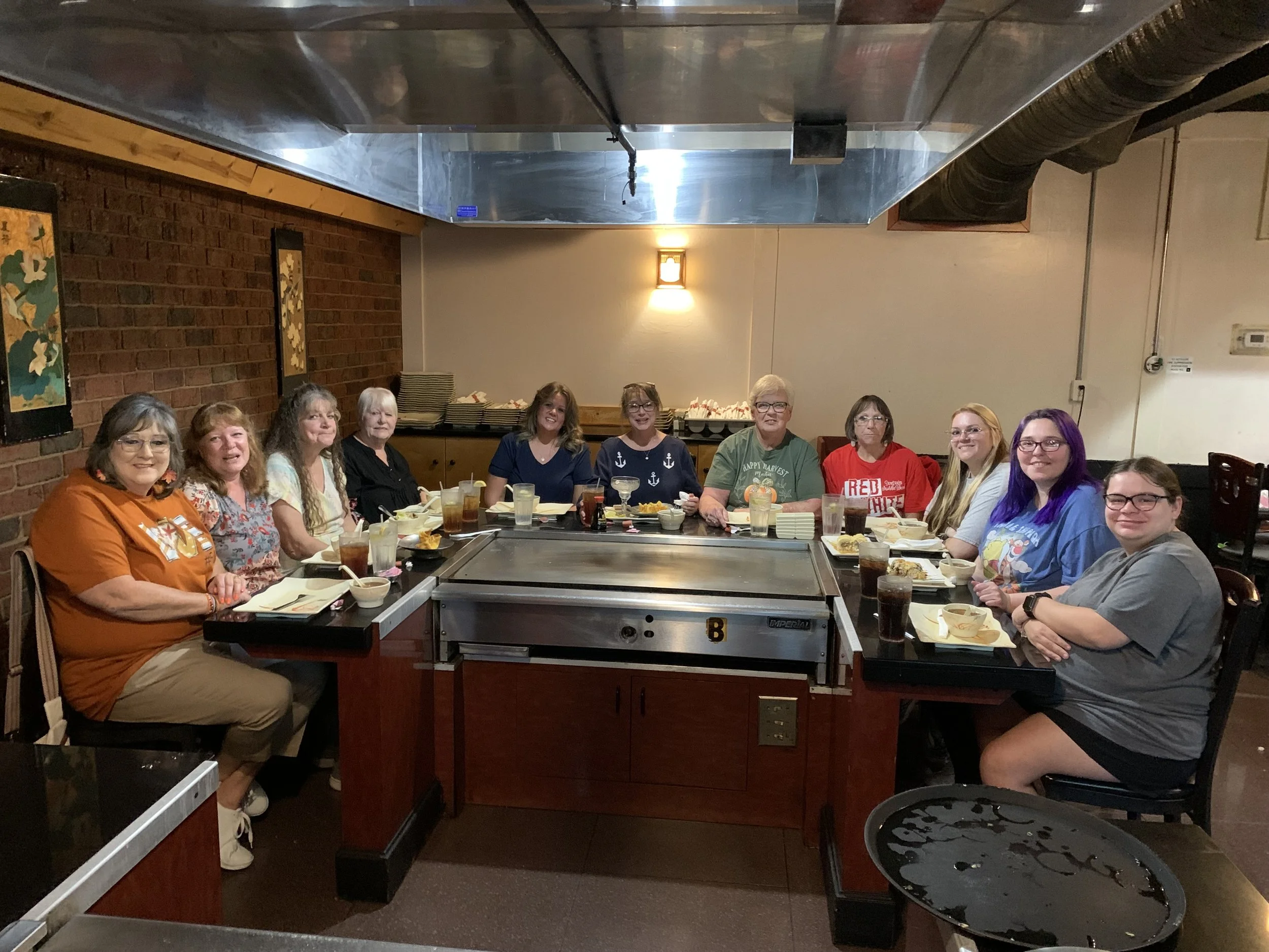 Group of people sitting around a rectangular table in a restaurant, smiling at the camera, with plates of food and drinks in front of them.