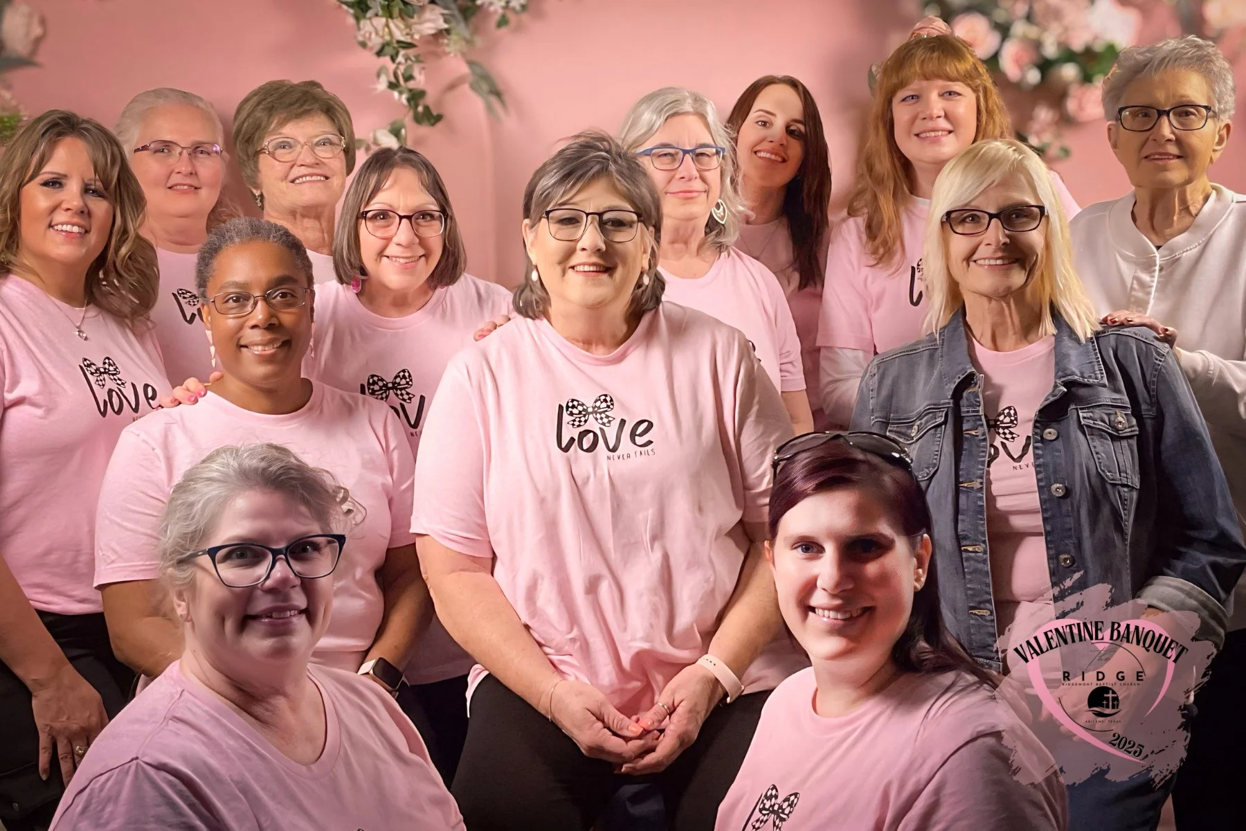 Group of women wearing pink T-shirts with the word love and a bow graphic, gathered for a photo at a Valentine's event in 2025, with pink and floral background.