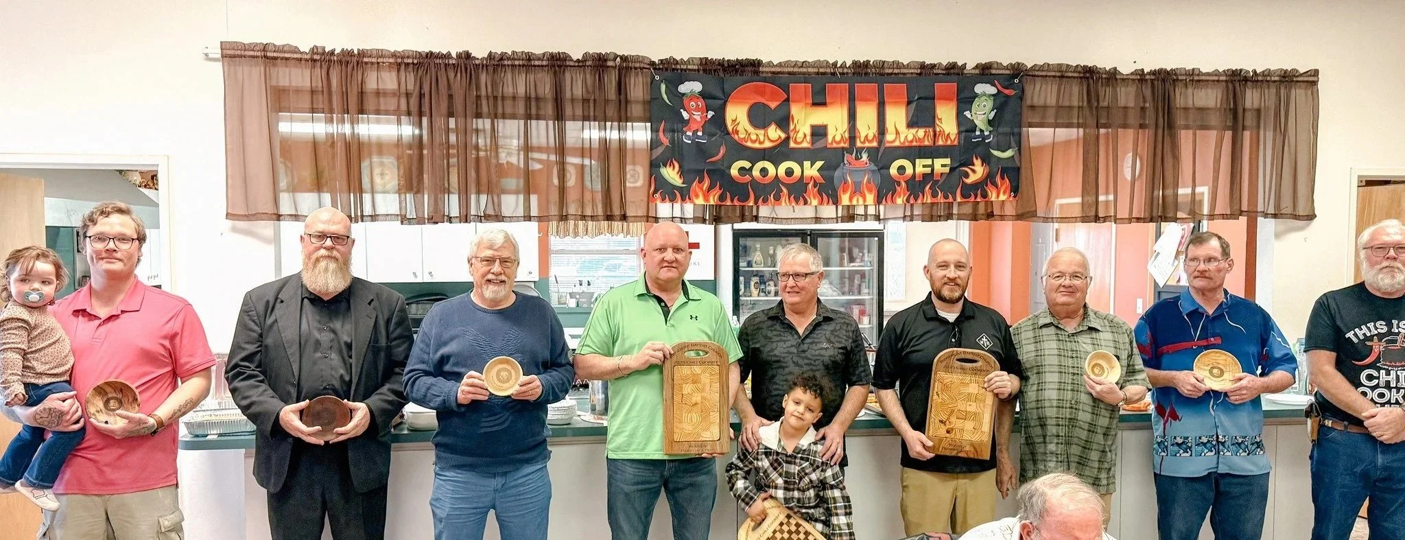 A group of nine men and two children standing in a room with a ‘Chili Cook Off’ banner hanging above them. Each person is holding a wooden serving tray or bowl, and they are gathered for a chili cook-off event.