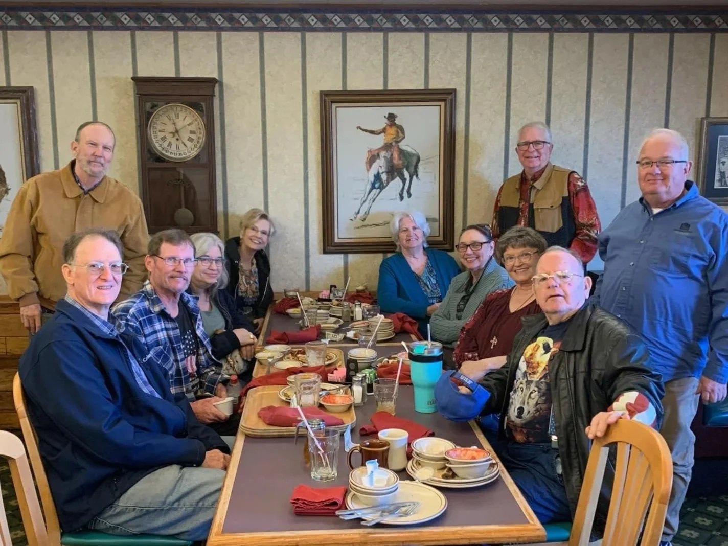 Group of eleven adults gathered around a restaurant table, enjoying a meal with plates of food, drinks, and napkins, in a room with painted wall art and a clock