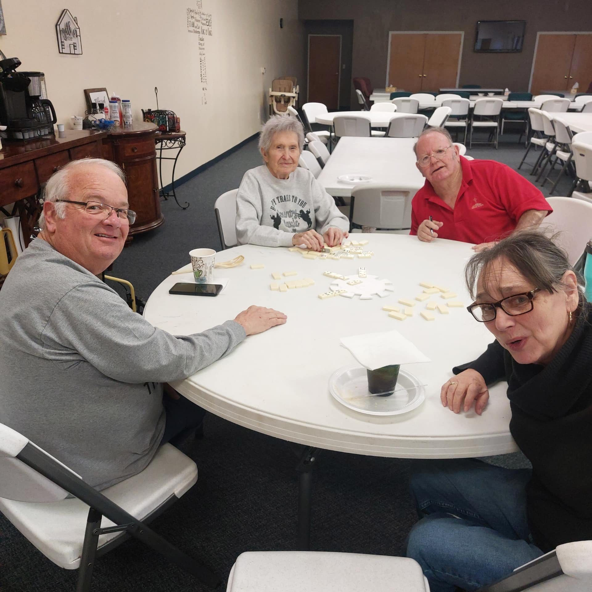 Four older adults sitting around a white oval table playing dominoes in a community room.