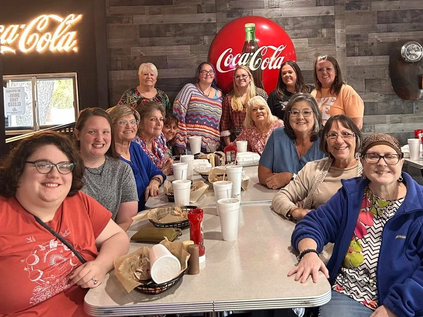 Group of women sitting and standing around a table in a restaurant, with Coca-Cola signs on the wall behind them.