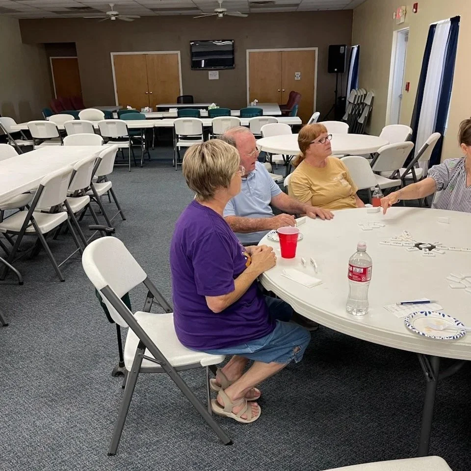 Three elderly people sitting at a large round table playing a game in a multi-purpose room with beige walls and foldable chairs.