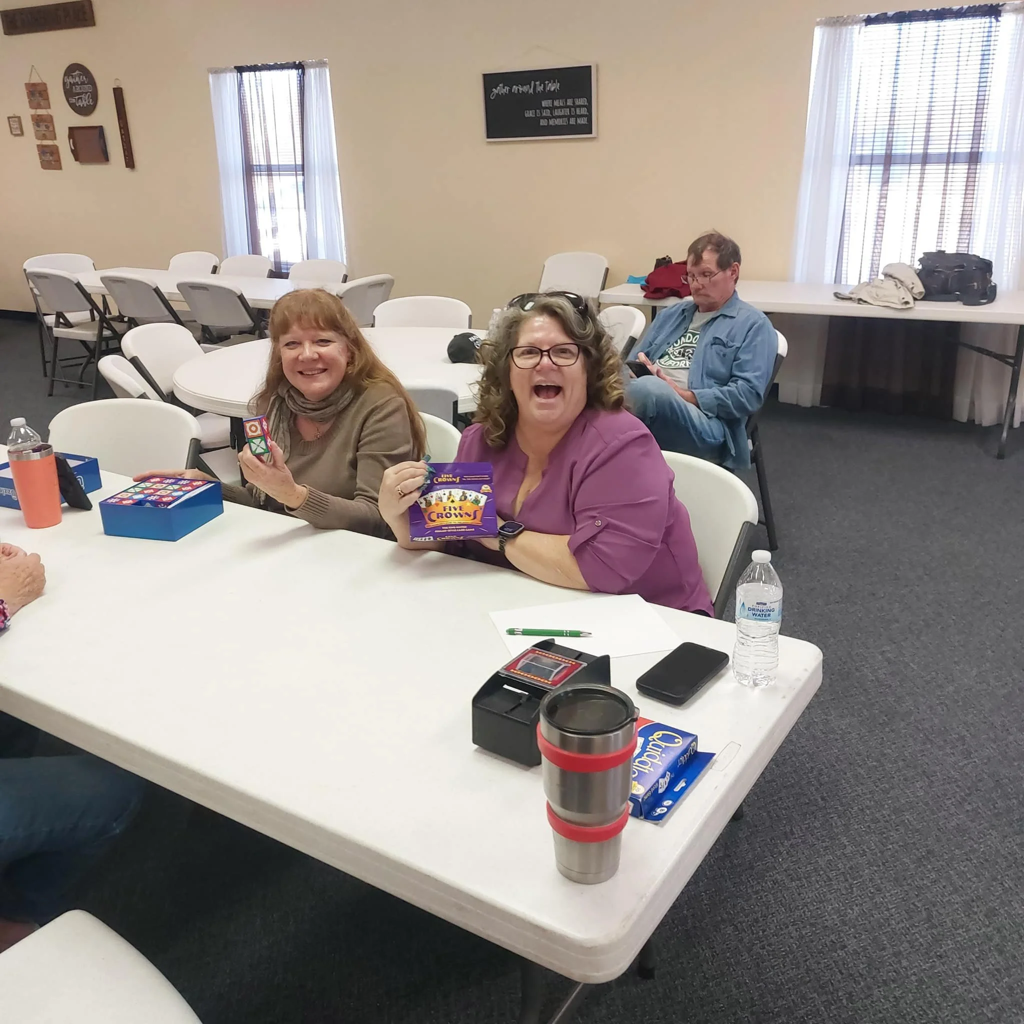 Three women sitting at a white table in a room with beige walls, holding small boxed games and smiling. Two women are in the foreground, one holding a game and laughing, the other holding a game box. A man is in the background using a smartphone. The table has items including a water bottle, a travel mug, a pen, and a small electronic device.