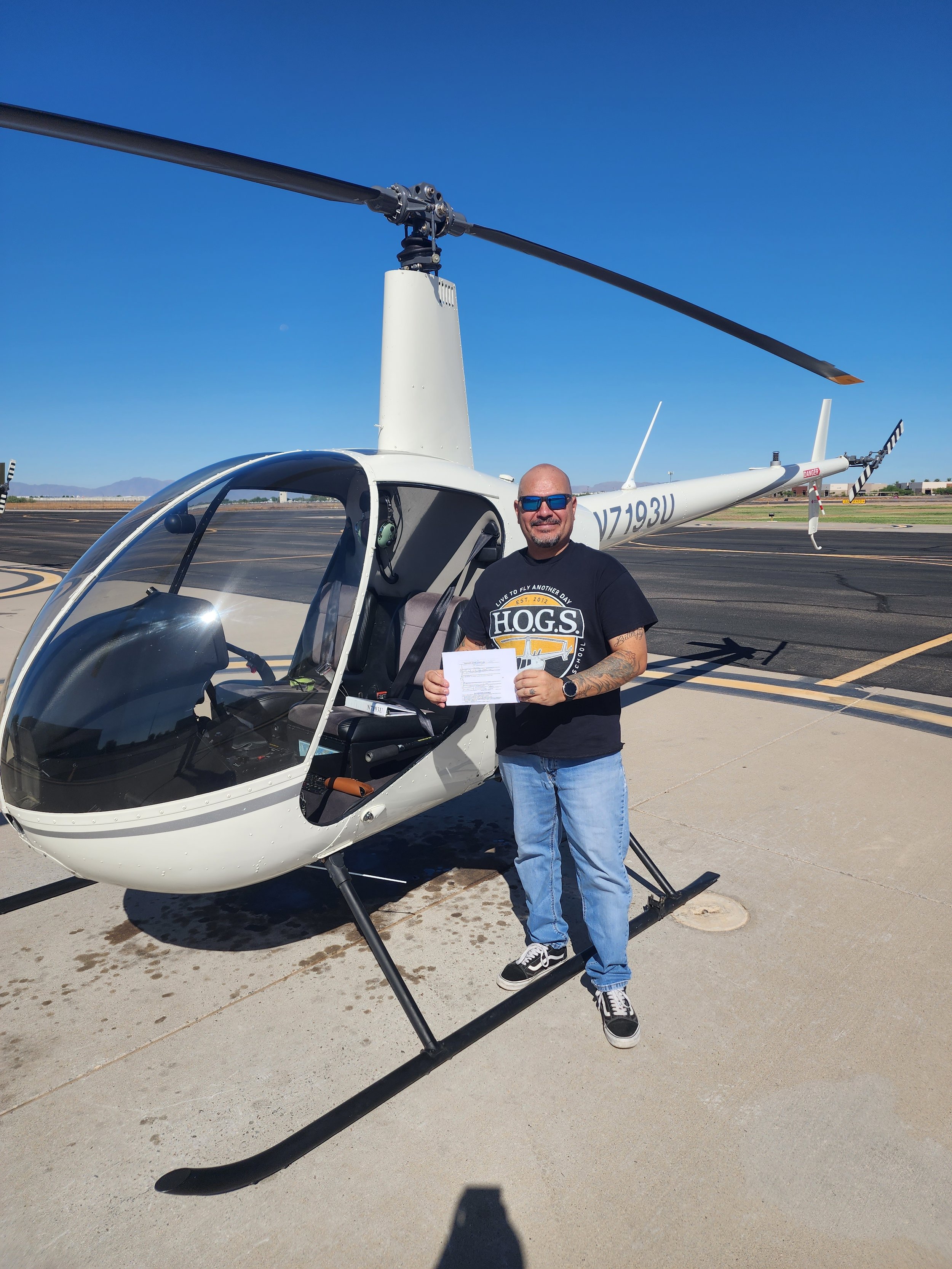 Man standing next to a small helicopter on an airport tarmac, holding a paper, wearing a black t-shirt, jeans, and sunglasses, with a clear blue sky in the background.