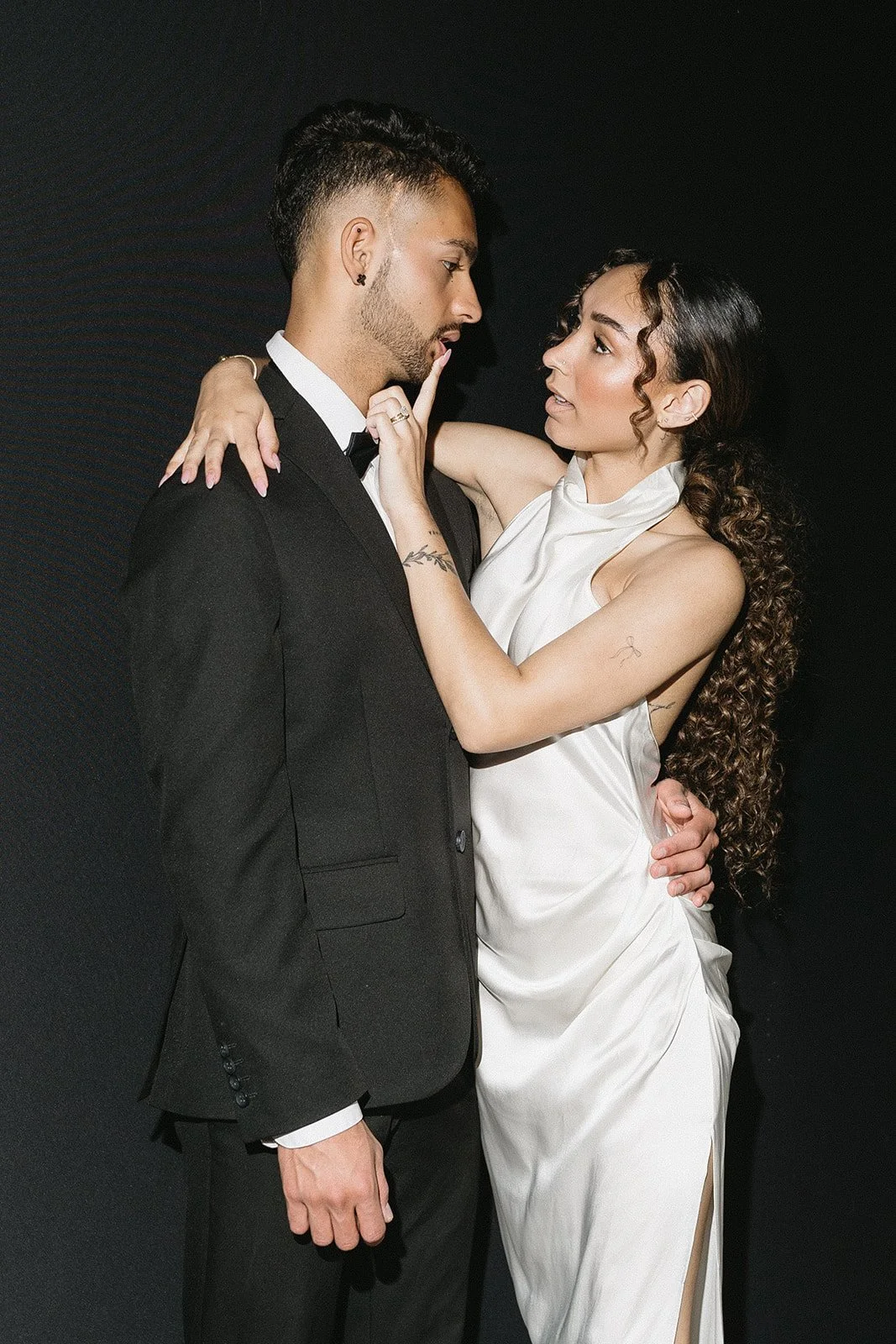 A man and woman dressed formally, standing close, with the woman gently touching the man's face and looking into his eyes against a black background.
