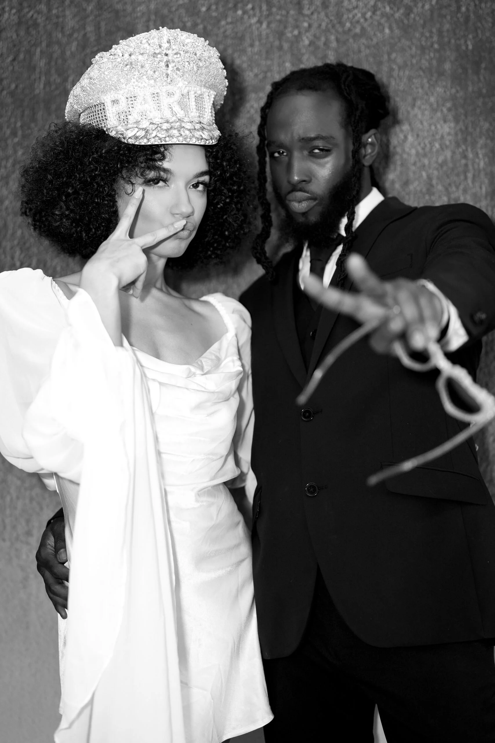 Two people posing for a photo, the woman wearing a hat with the word 'PARTY' and making a peace sign, the man wearing a suit and making a peace sign with his hand.