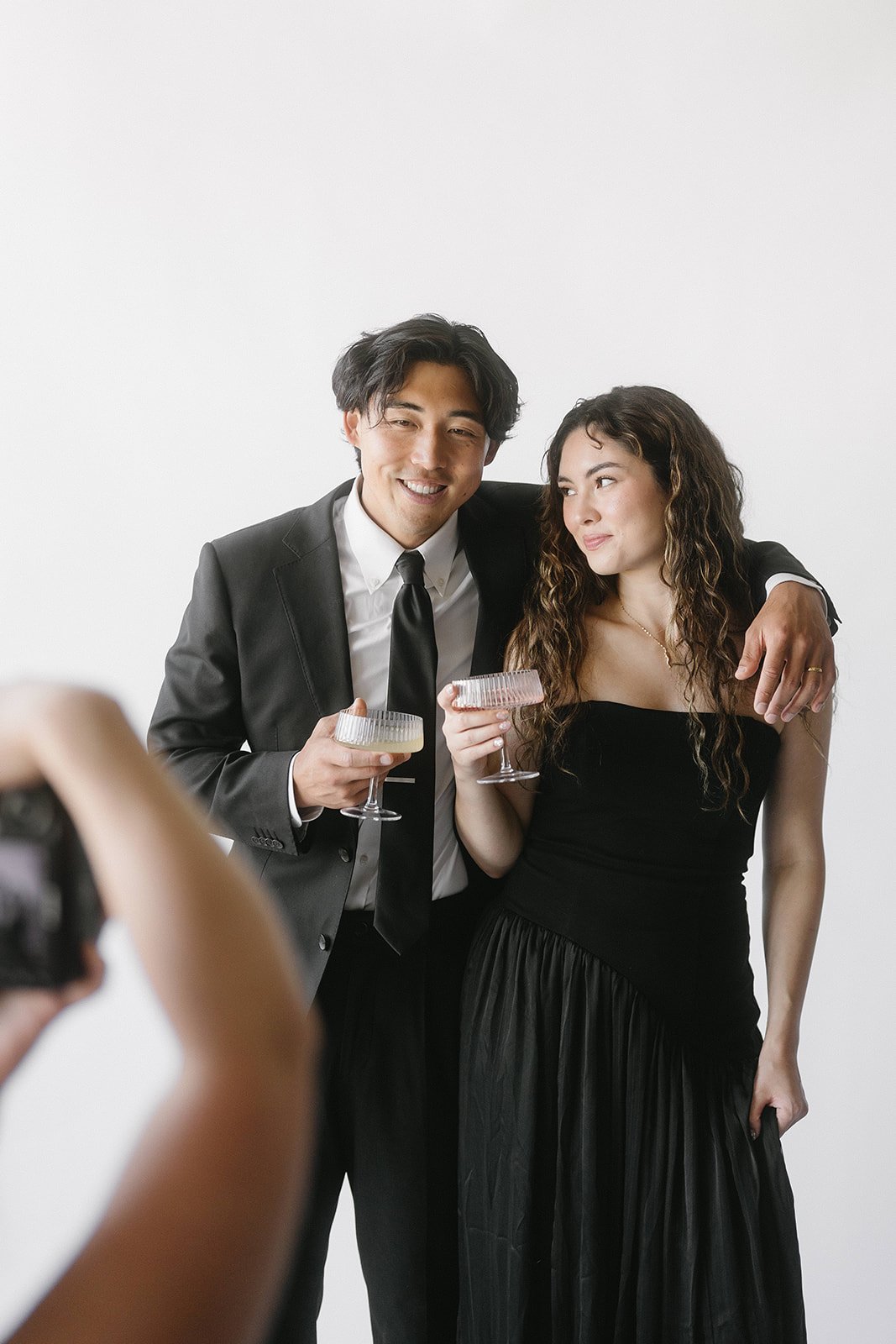 A couple dressed in formal attire, the man in a suit and tie and the woman in a black dress, pose together with drinks in a photo studio.