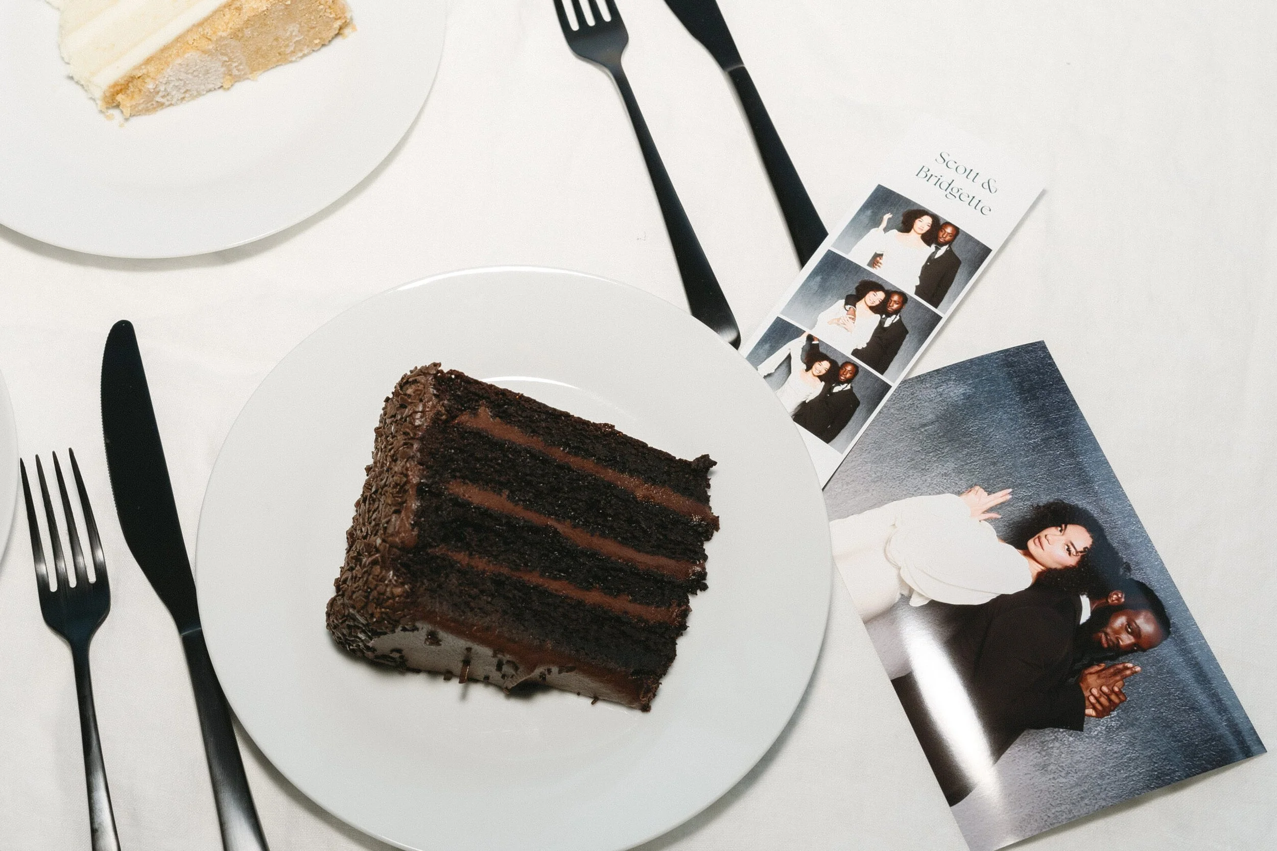 A slice of chocolate layered cake on a white plate, adjacent to another plate with a slice of vanilla cake, on a white table. There are black utensils arranged around the plates, and a photo strip with three pictures of a couple, along with a separate printed photograph of the same couple in a different pose.