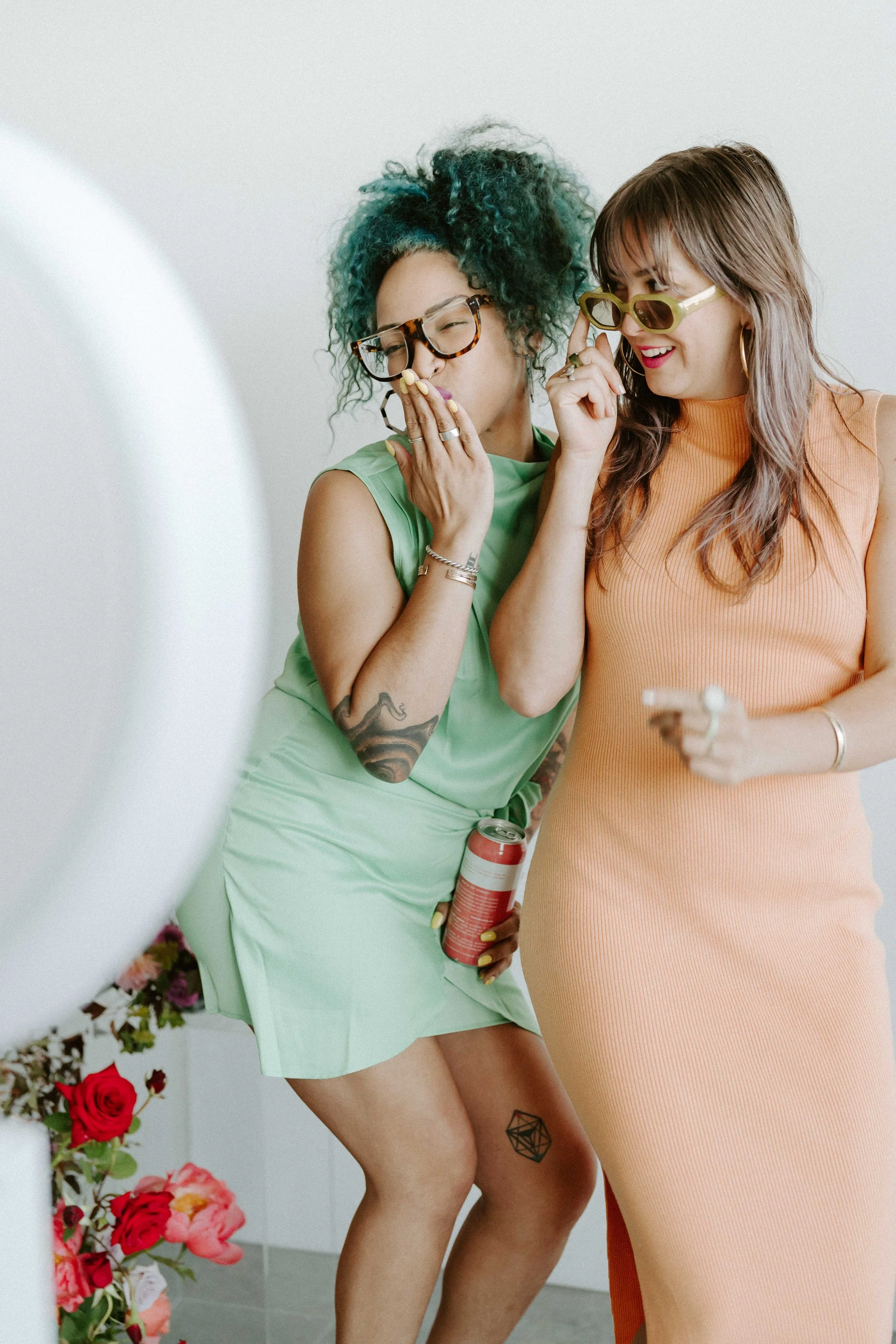Two women in colorful dresses posing playfully, one holding a soda can, with flowers visible in the foreground.
