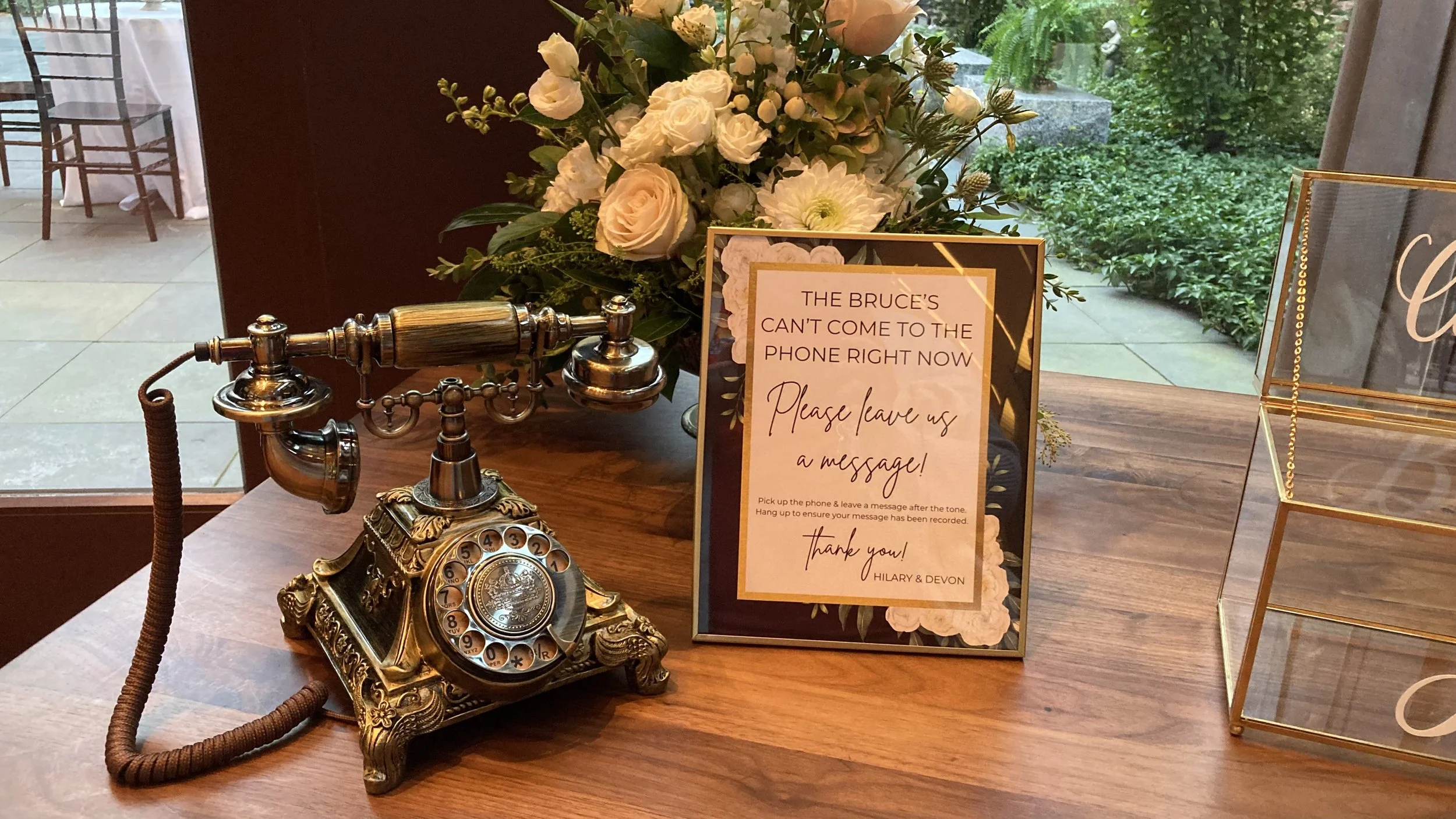 An antique brass rotary phone on a wooden table next to a sign and a floral arrangement. The sign reads 'The Bruce's can't come to the phone right now. Please leave us a message!' and instructs to pick up the phone and leave a message after the tone.