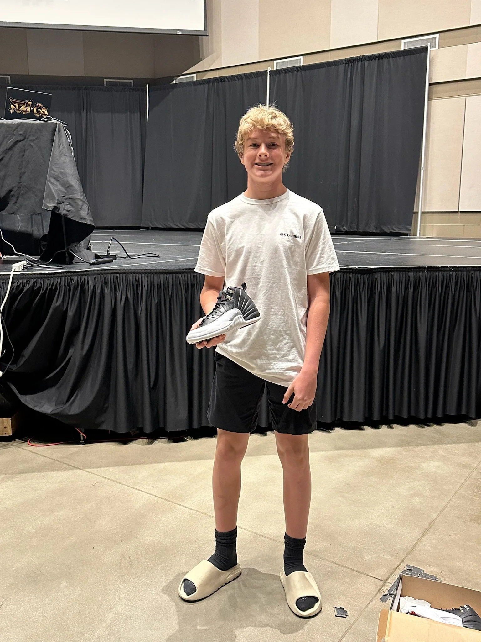 A young man standing indoors on a stage holding a sneaker, wearing a white t-shirt, black shorts, black socks, and slide sandals, with a black curtain in the background.