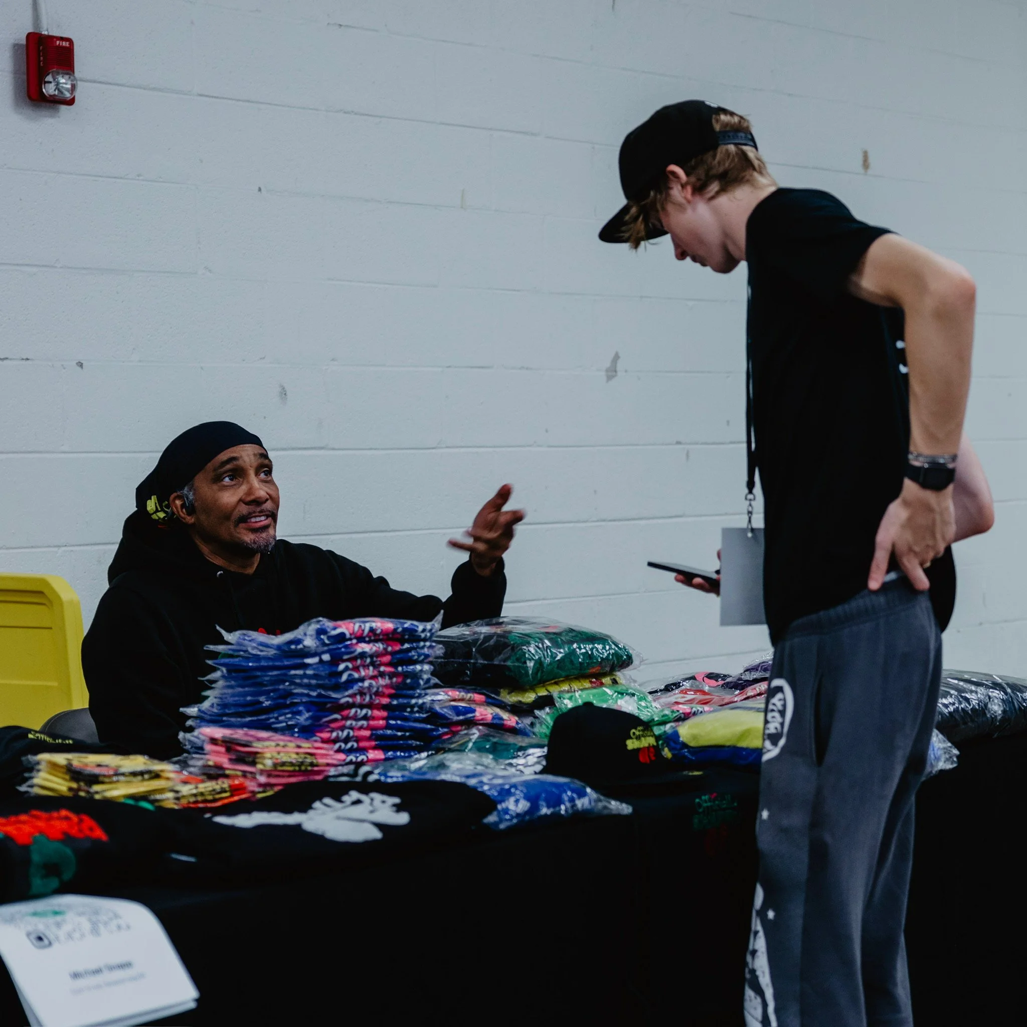 A man wearing a black hoodie and head covering sitting behind a table with various clothing items, talking to a young man in a black t-shirt and gray sweatpants, in an indoor setting with a white brick wall.