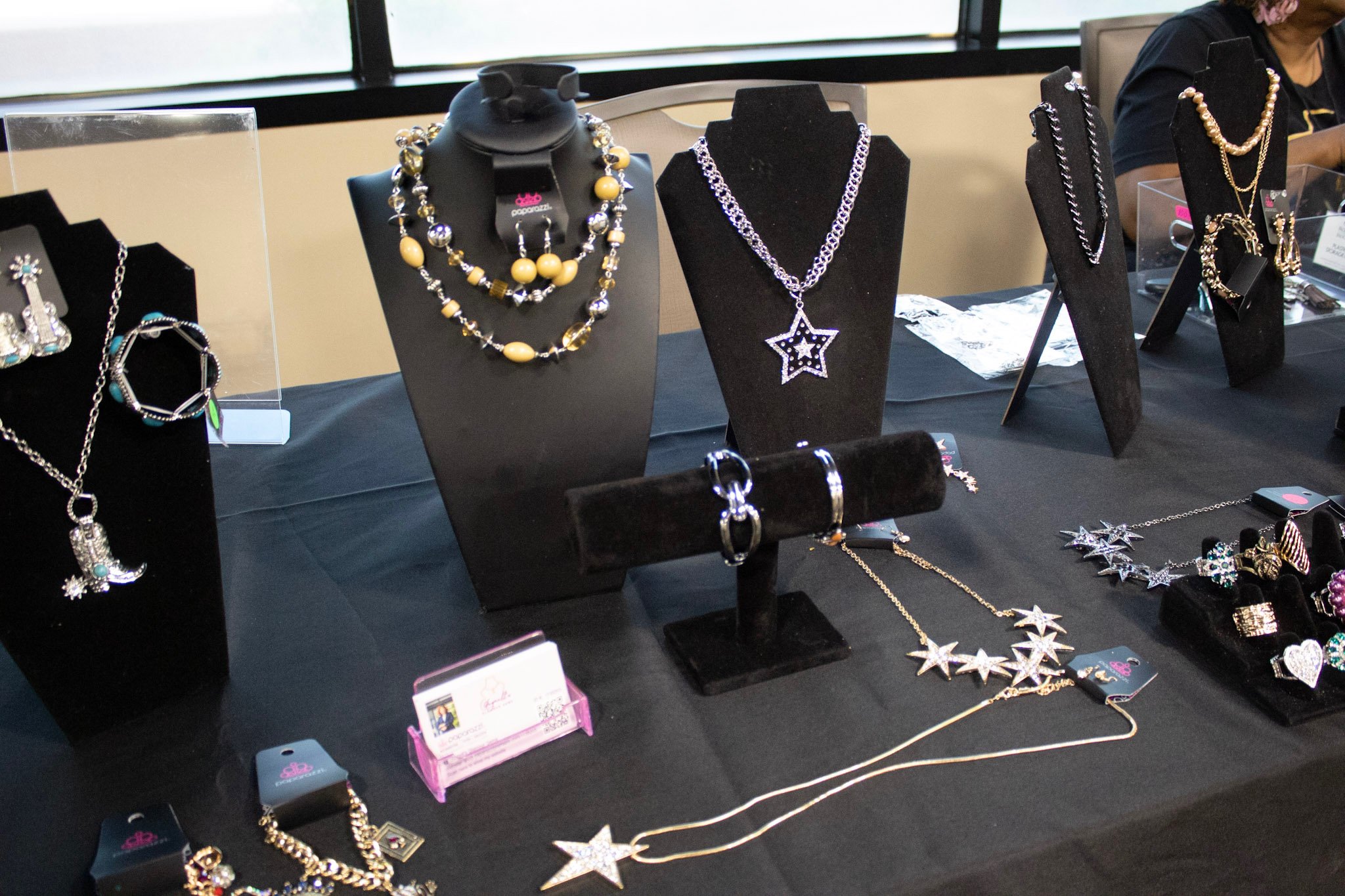 Jewelry display table with necklaces, bracelets, and rings, including star and moon themed pieces, at an indoor market or craft fair.