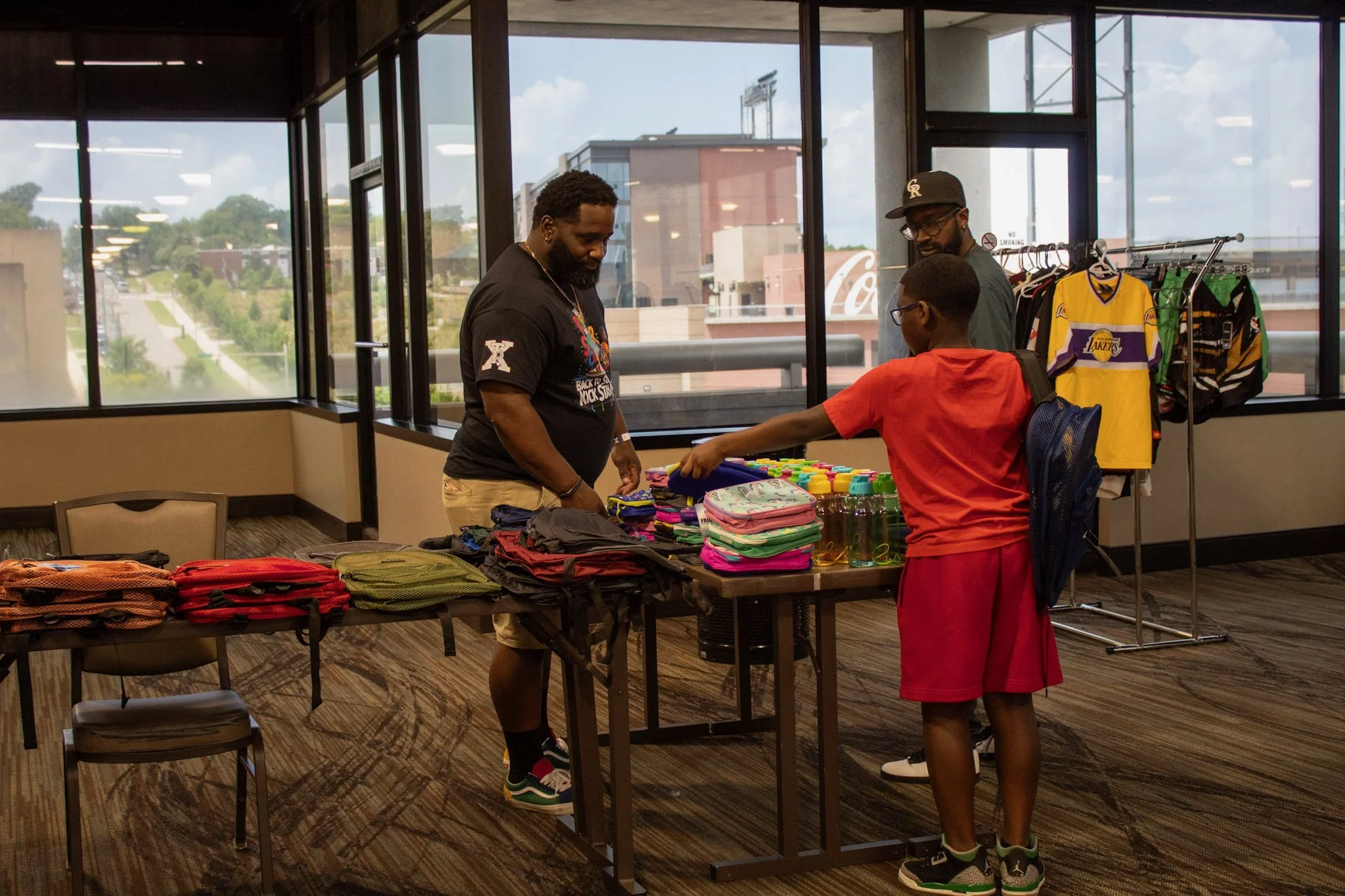 Three people looking at backpacks and water bottles at a table near large windows with a cityscape view.