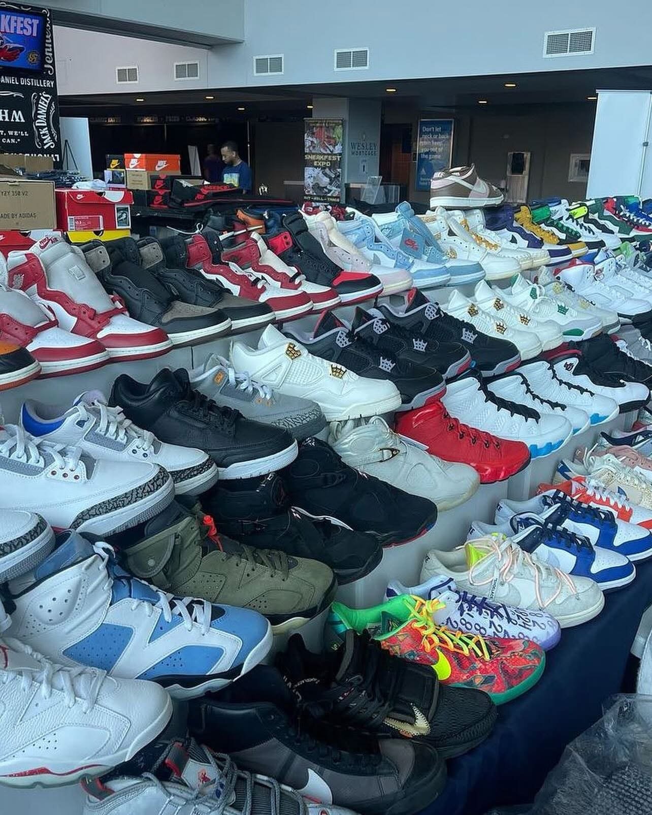 Table filled with various new Nike sneakers, with multiple pairs of athletic shoes in different colors and styles displayed at an indoor event or sale.
