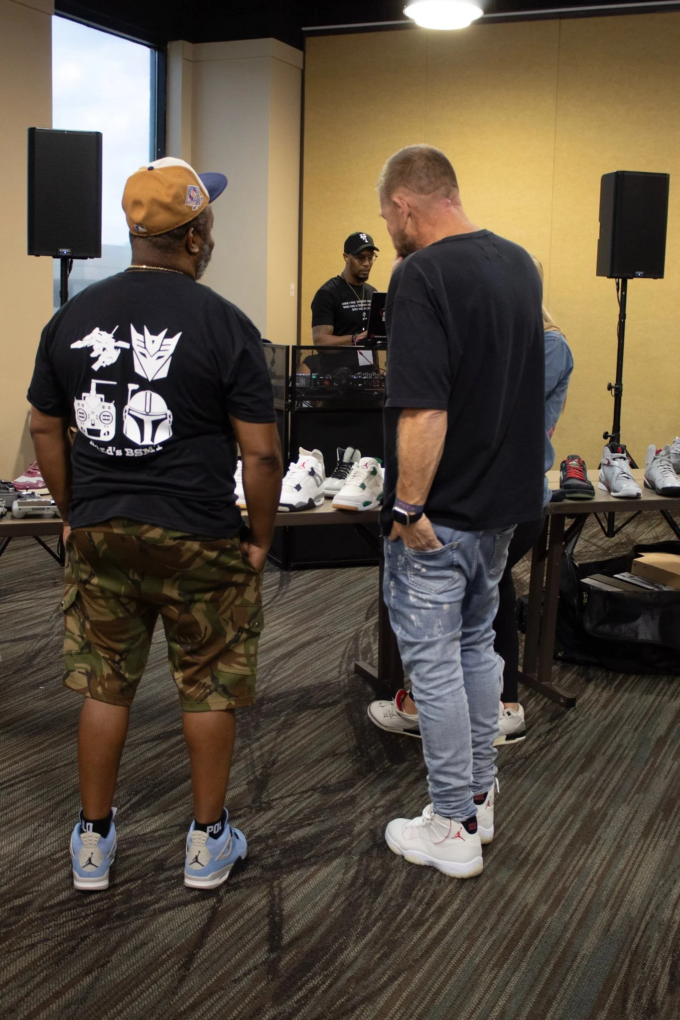Two men standing and talking in front of a table with sneakers, with a DJ playing music in the background at an indoor sneaker event.