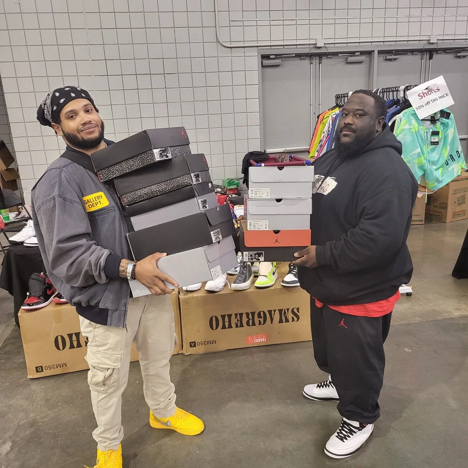Two men holding shoe boxes at a thrift or consignment store with sneakers and clothing on display behind them.