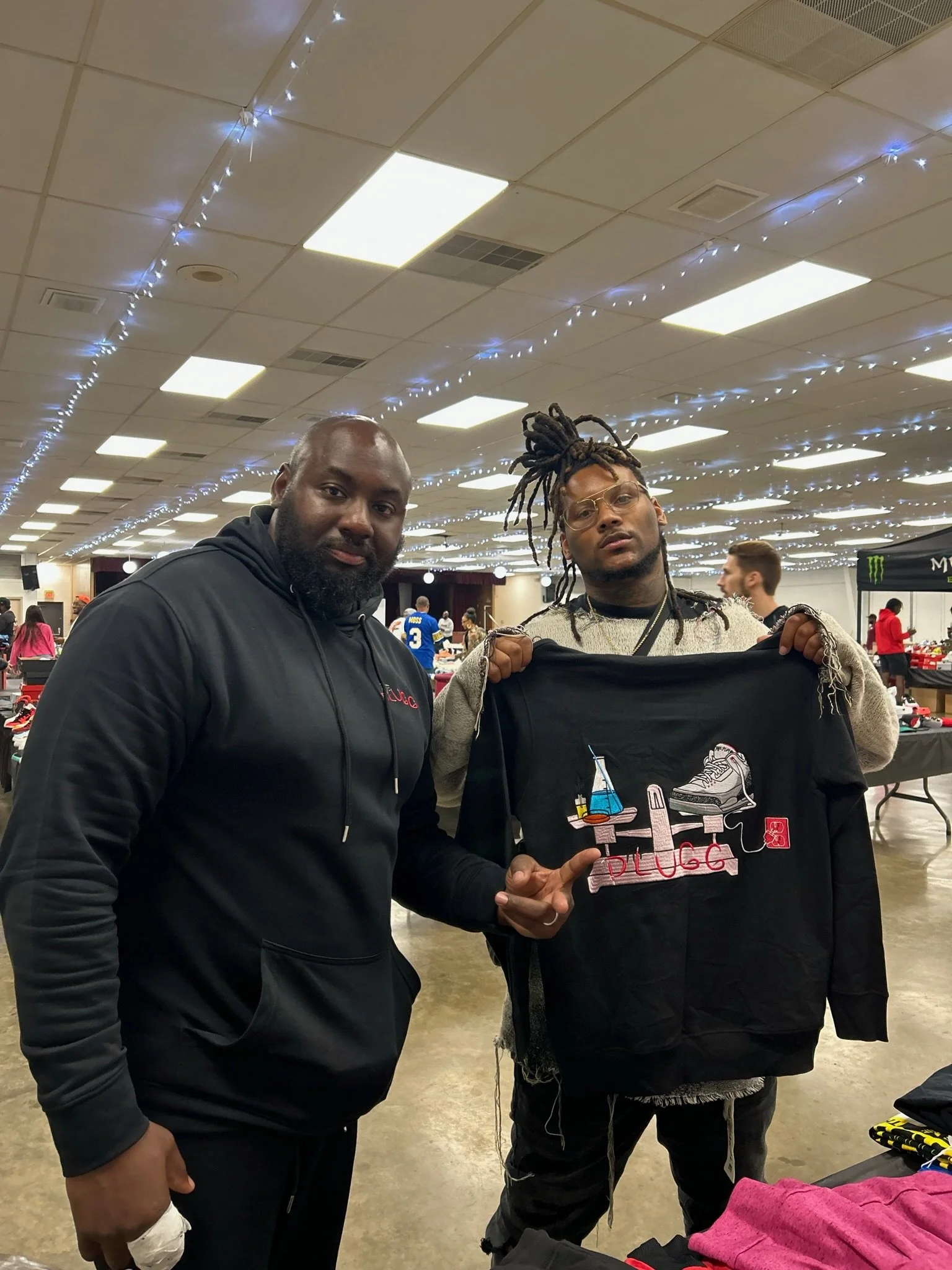 Two men in an indoor event space, one holding a black jacket with a colorful embroidery logo and text, other pointing at it, with tables and other people in the background.
