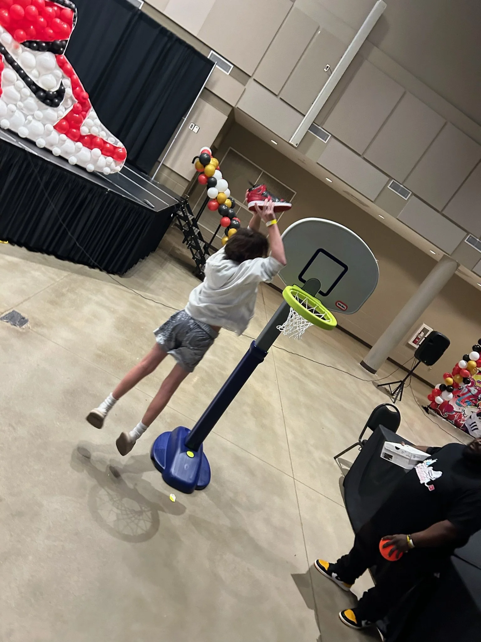 A person jumping to shoot a toy basketball into a small indoor basketball hoop at an event with holiday decorations, including a balloon sculpture of Santa Claus, in the background.