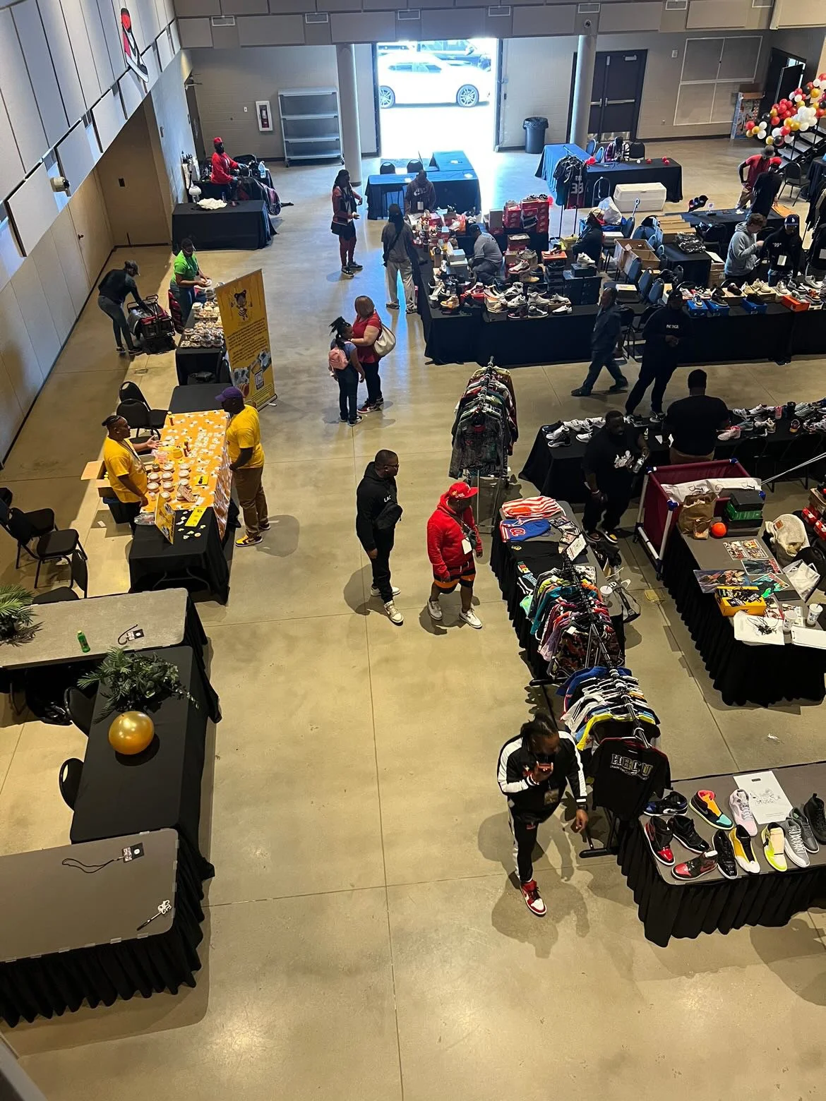 Indoor thrift or flea market with tables displaying shoes, clothing, and various items, and people shopping and browsing.