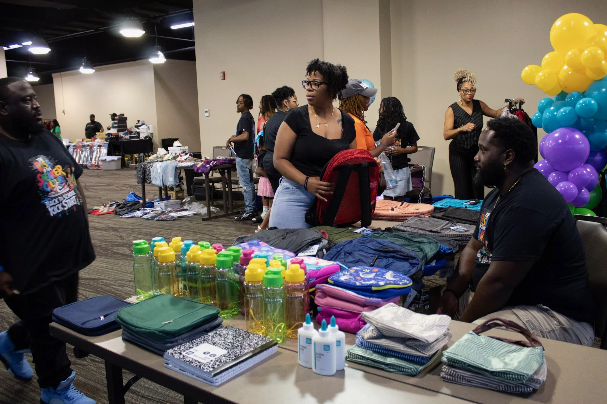 People shopping at a vendor booth with water bottles, backpacks, notebooks, and fabric on tables, with balloons in rainbow colors in the background.