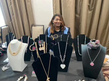 Woman standing behind a jewelry display table with various necklaces and jewelry pieces.