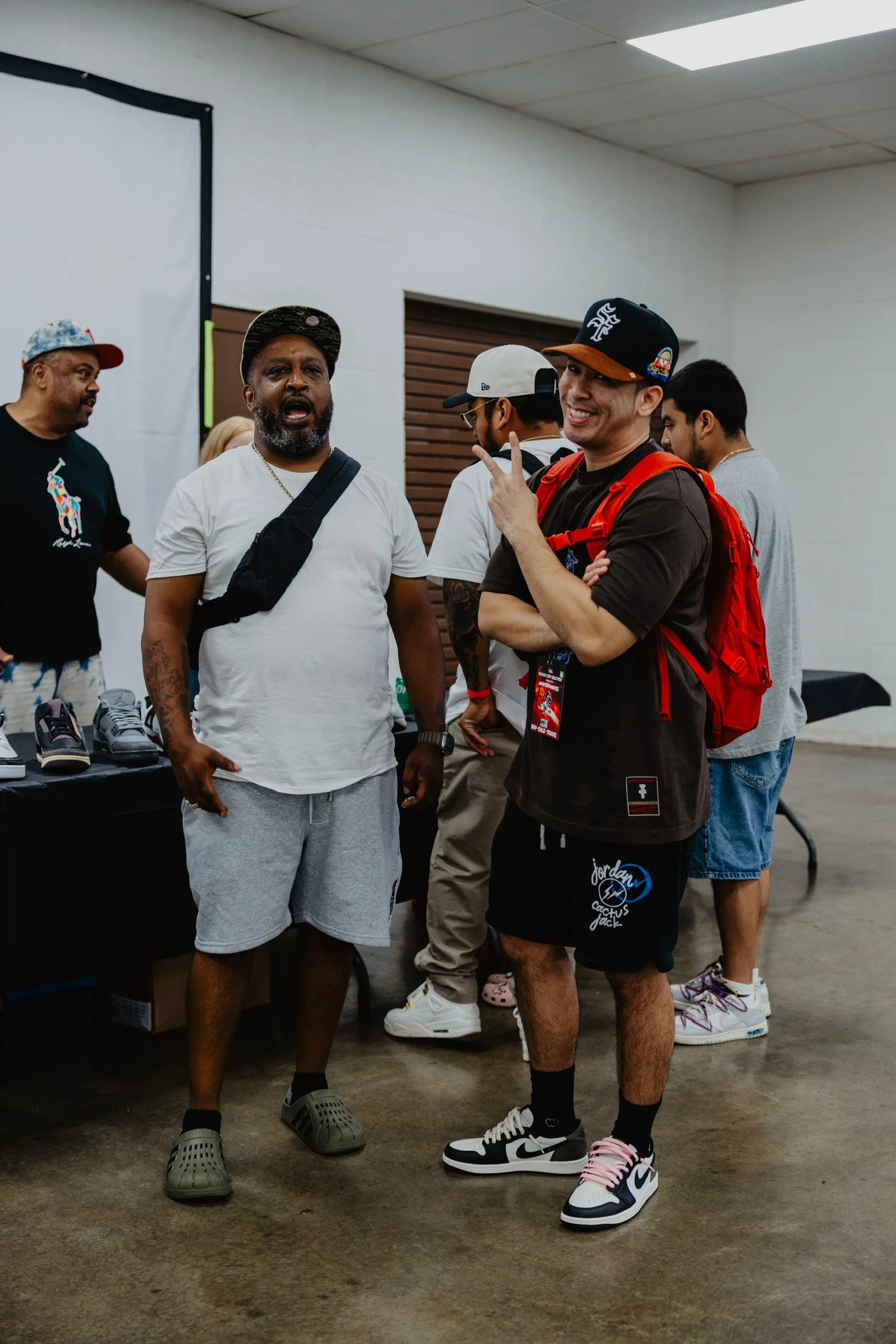 Group of men chatting indoors, with some wearing casual sportswear and sneakers, and a table with shoes in the background.