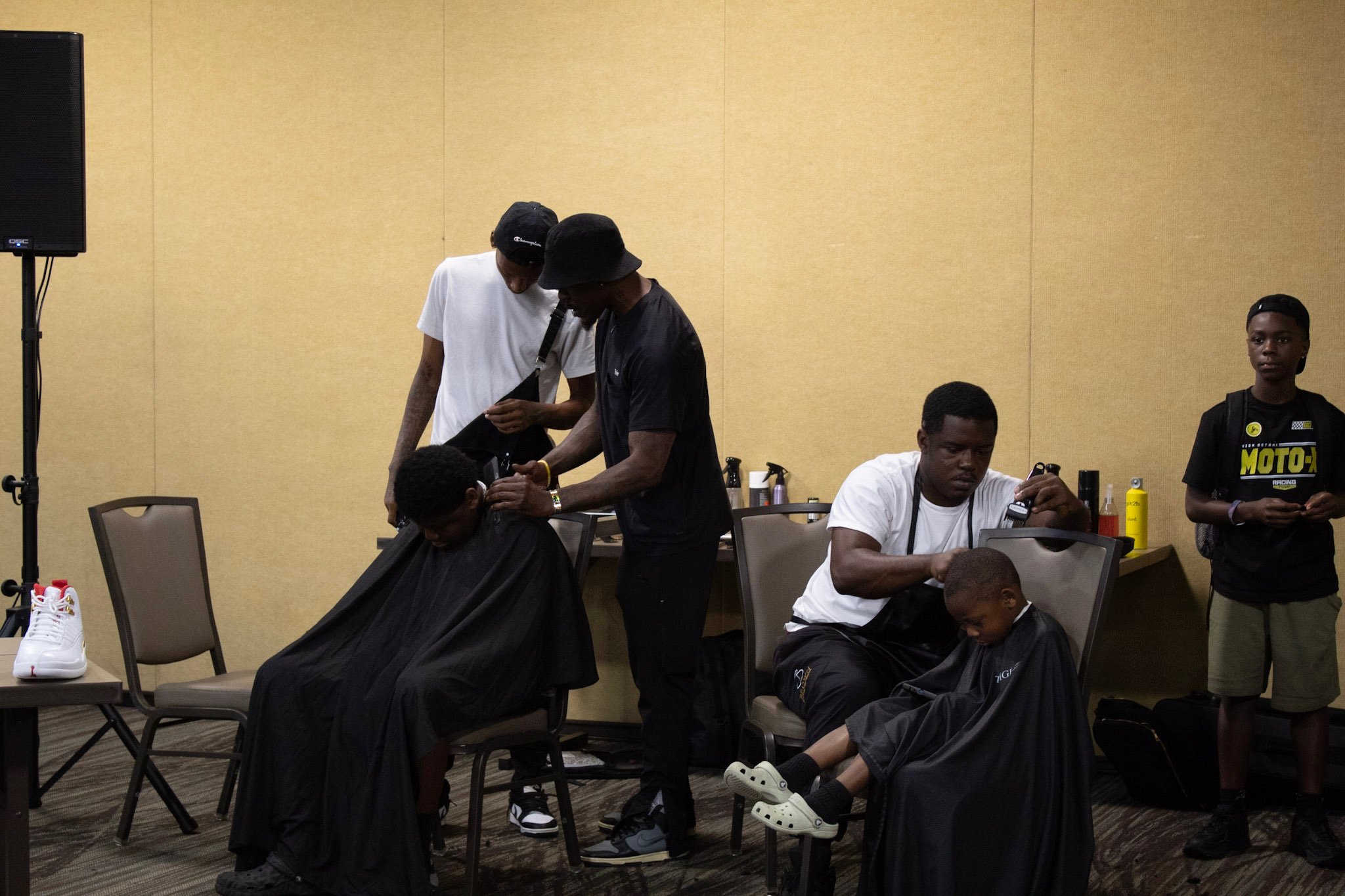 Two men give haircuts to young boys in a casual indoor setting, with others standing nearby and various supplies on the countertop.