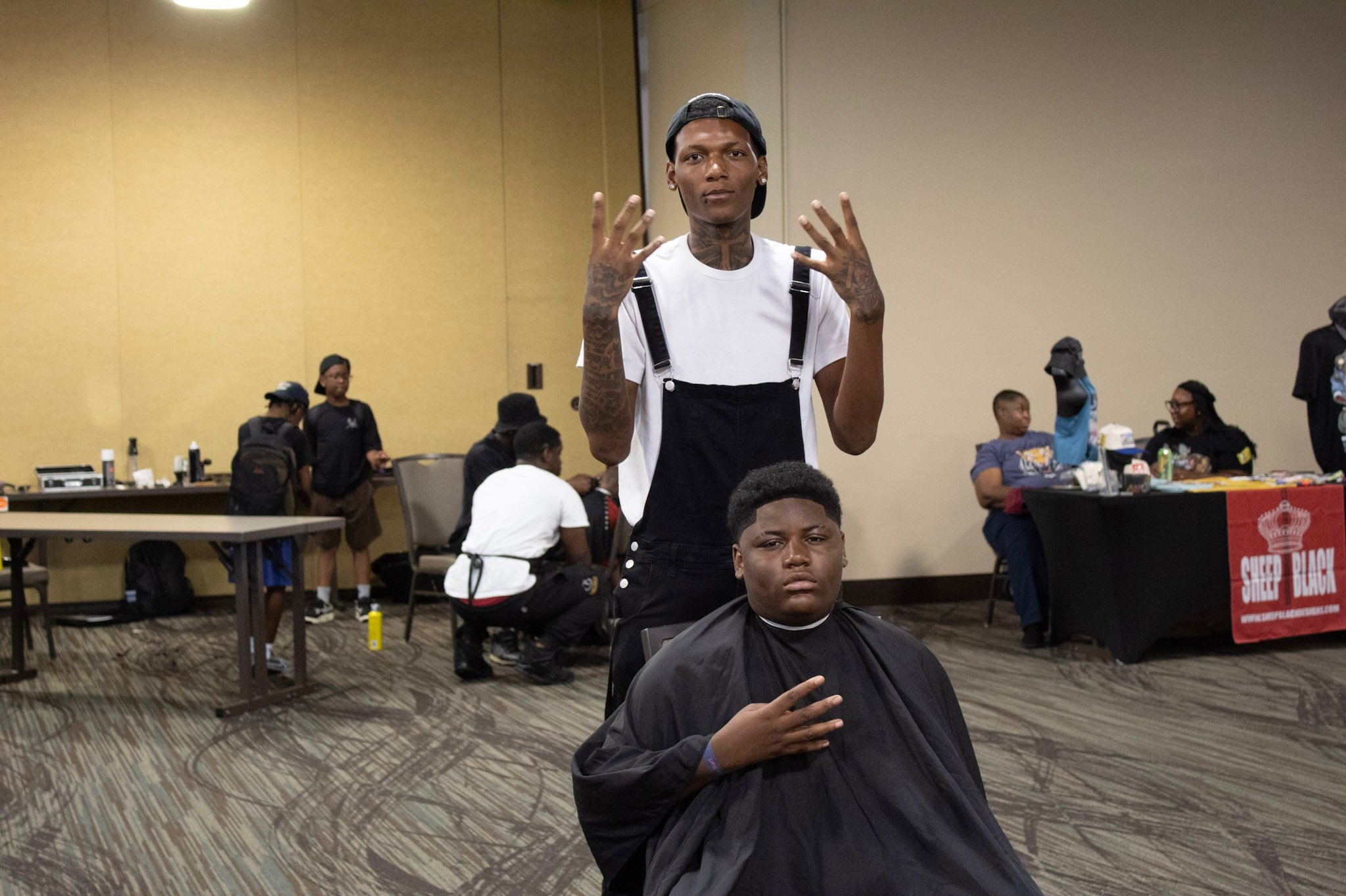 A young man with tattoos, wearing a white t-shirt, black overalls, and a backwards baseball cap, stands behind another young man seated in a barber's cape. The seated young man has a serious expression. In the background, there are people, tables, an