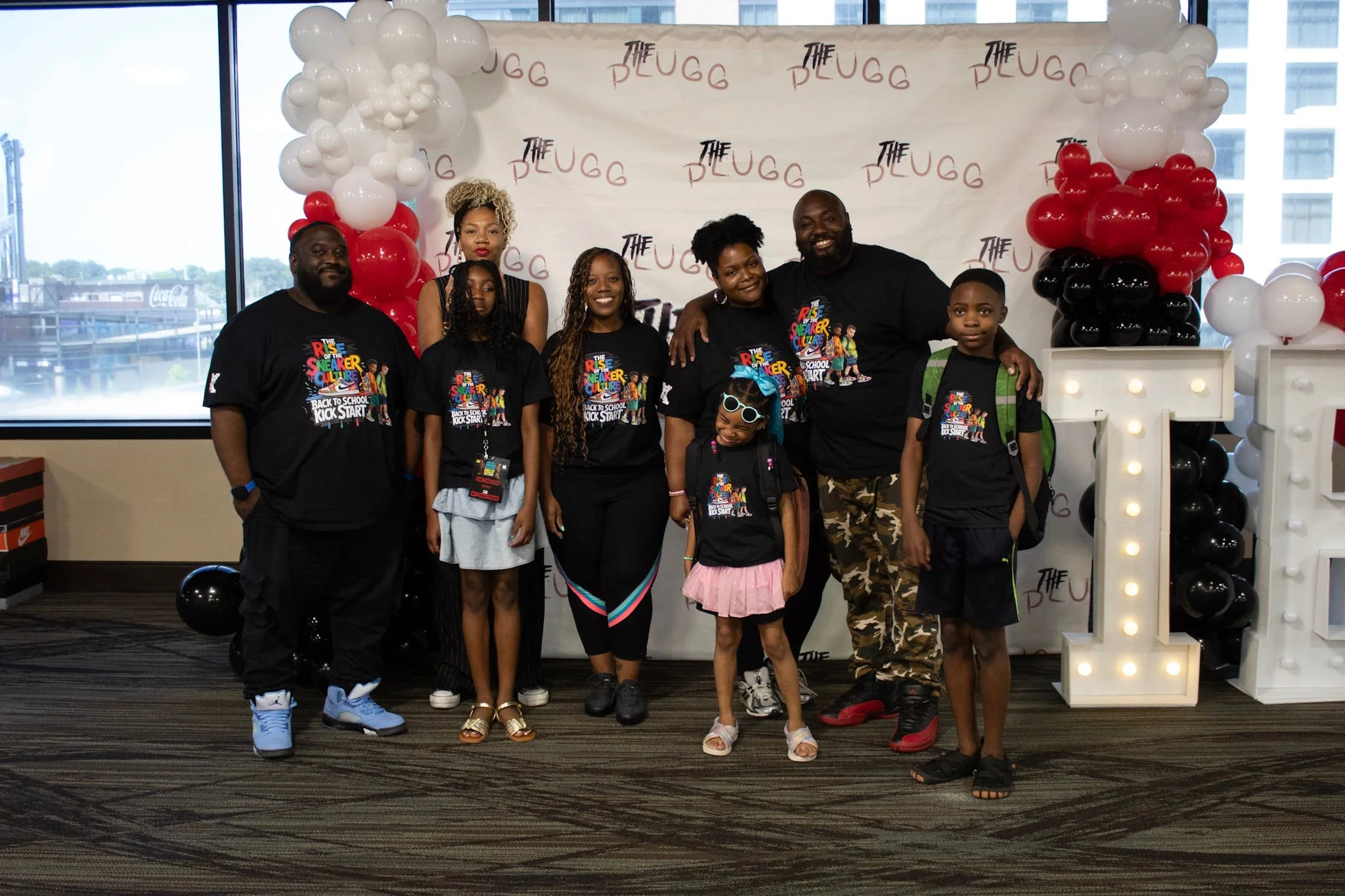 Group of eight people, including children and adults, standing in front of a backdrop with balloons and event branding. All are wearing black T-shirts with colorful printing that reads 'The Rise of the Sneaker Culture' and 'Back to School Kick Start.