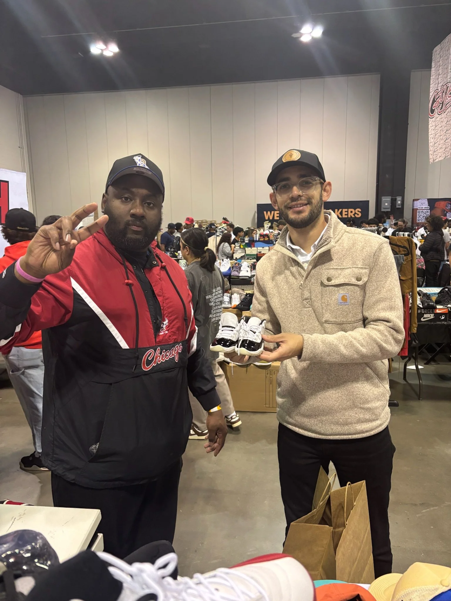 Two men standing inside a sneaker event, with tables of shoes behind them. The man on the left is wearing a black and red Chicago sports jacket and a black cap, making a peace sign. The man on the right is wearing a beige Carhartt jacket, glasses, an