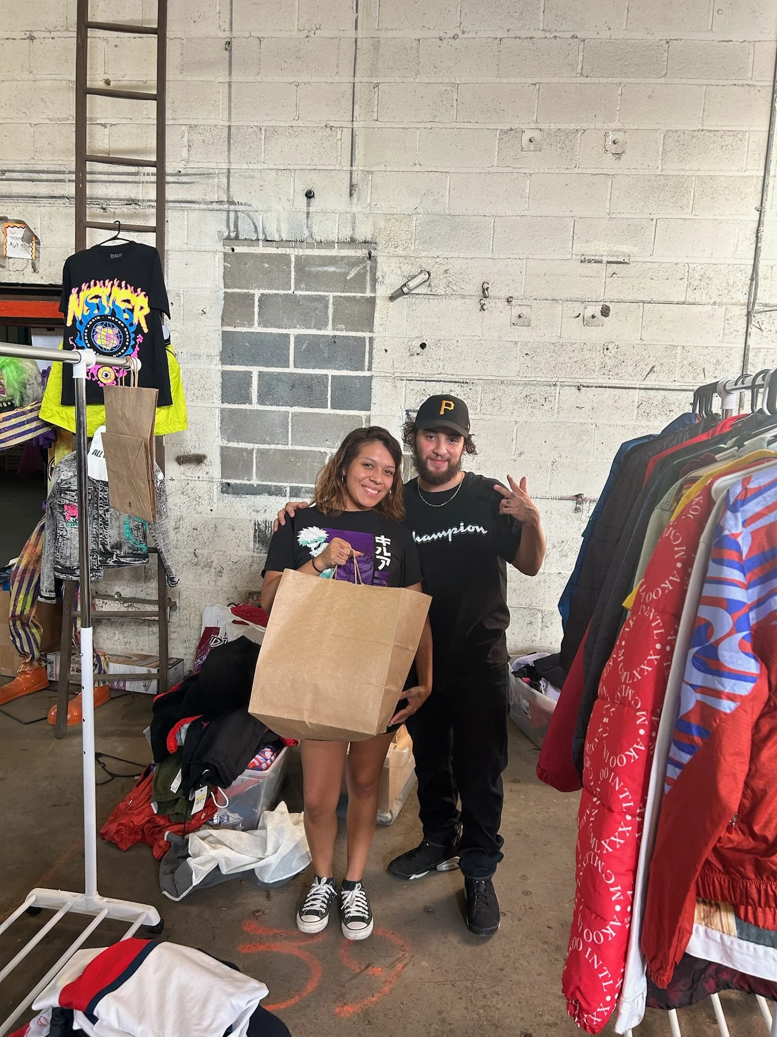A woman and a man standing inside a thrift store, smiling at the camera. The woman is holding a large paper shopping bag, wearing casual clothing and sneakers. The man is making a peace sign with his right hand, wearing a black cap, black t-shirt, an
