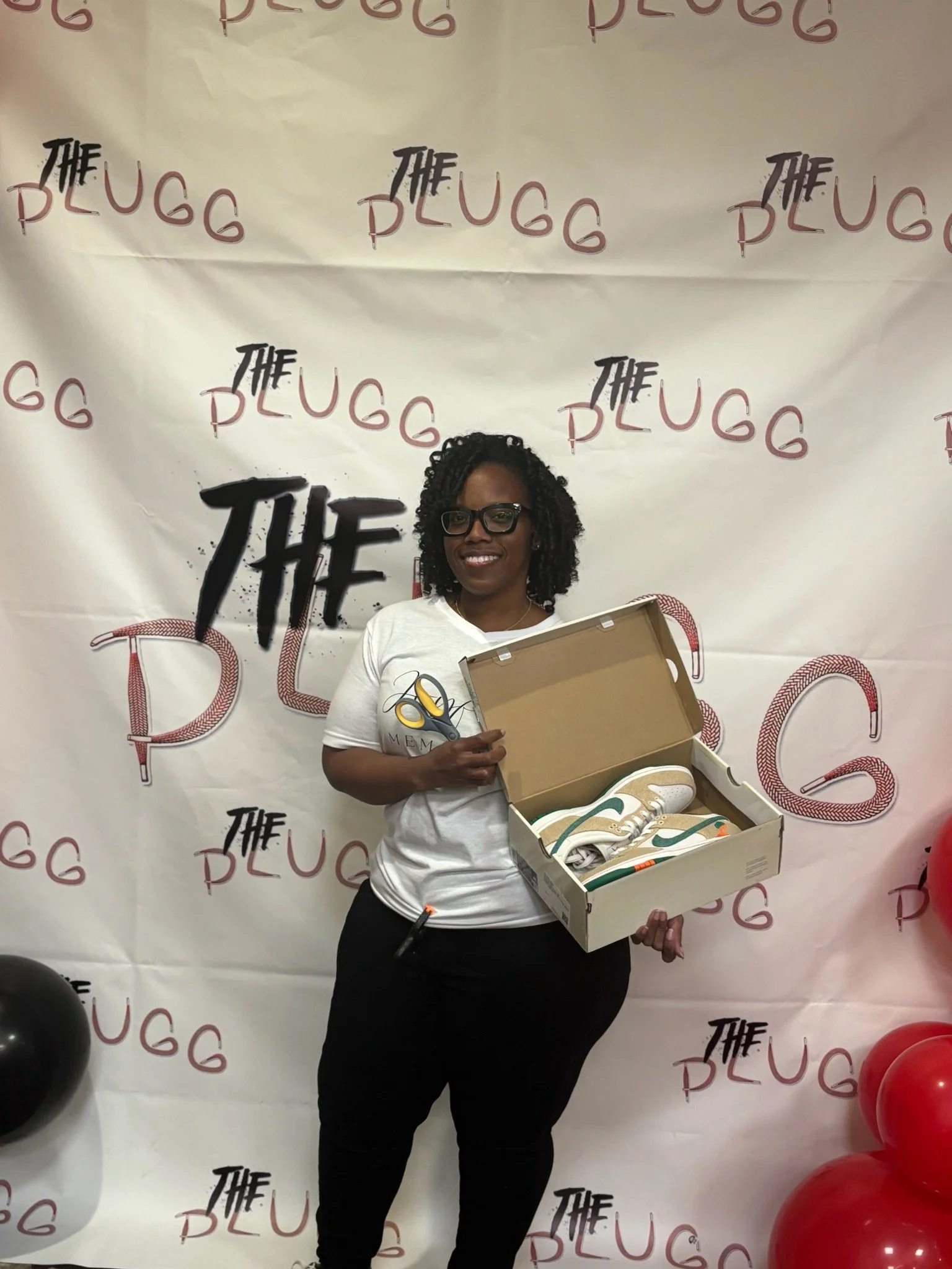Woman with curly hair and glasses smiling and holding a box of sneakers in front of a backdrop with 'The Pugg' logo, red and black balloons on the side.