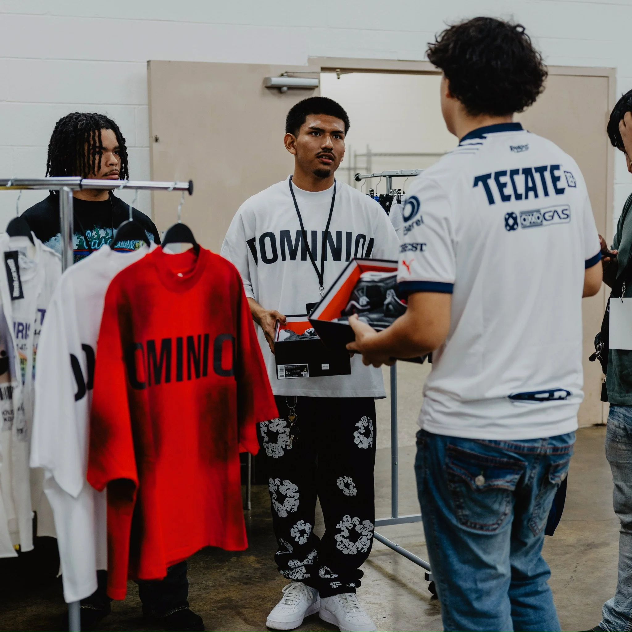 Three young men in a room with clothing racks and shirts on display, engaged in conversation about sports or fashion, with one holding a box of shoes.