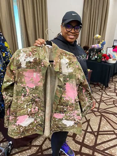 A smiling woman wearing glasses, a black Nike cap, and a dark hoodie, holding a large camouflage jacket with pink and white paint splatters in an indoor setting with beige curtains and a table with decorative items in the background.