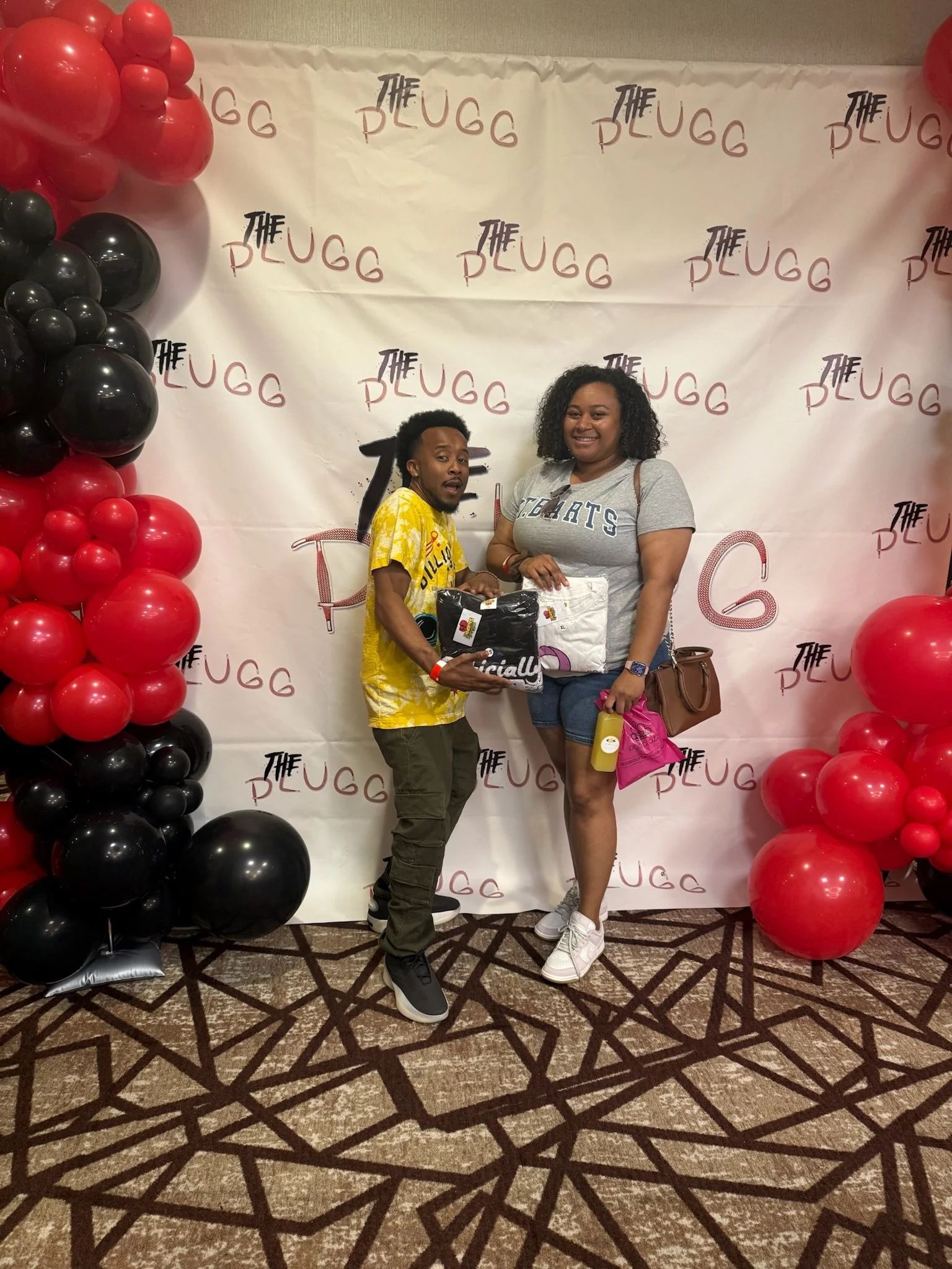A man and woman standing together in front of a backdrop with red and black balloons on either side and the words 'The PLUGG' repeated on it. The man is wearing a yellow shirt and dark pants, holding a package, while the woman is wearing a gray t-shi
