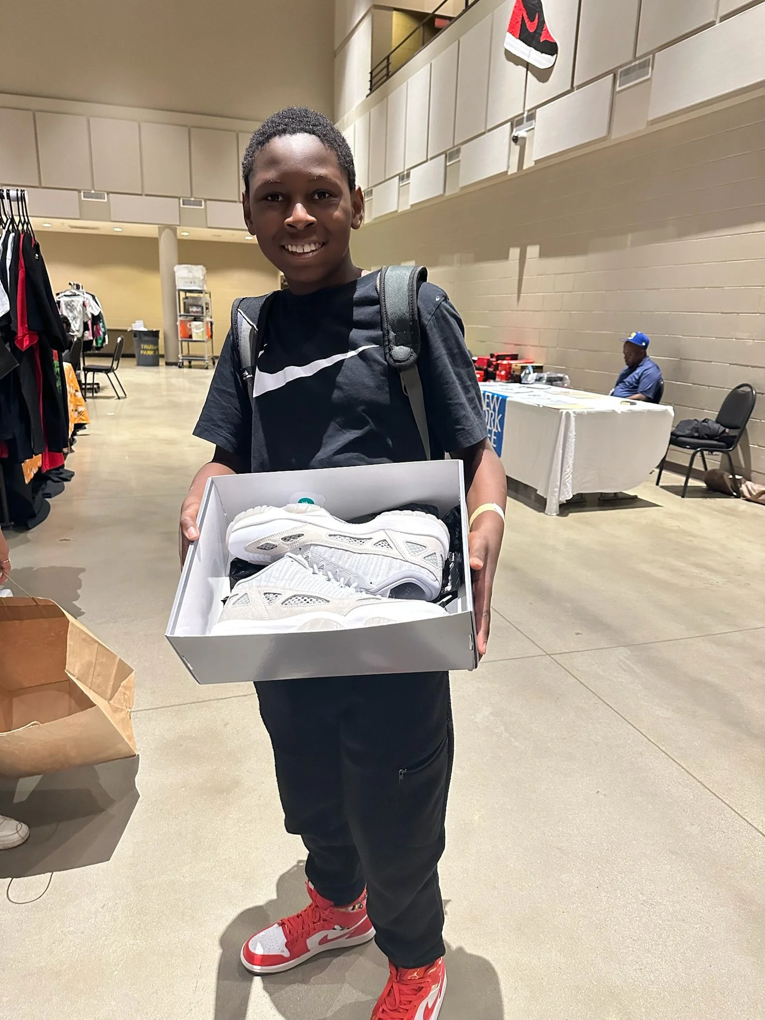 A smiling boy with a backpack holding a box of white athletic shoes at an indoor event.