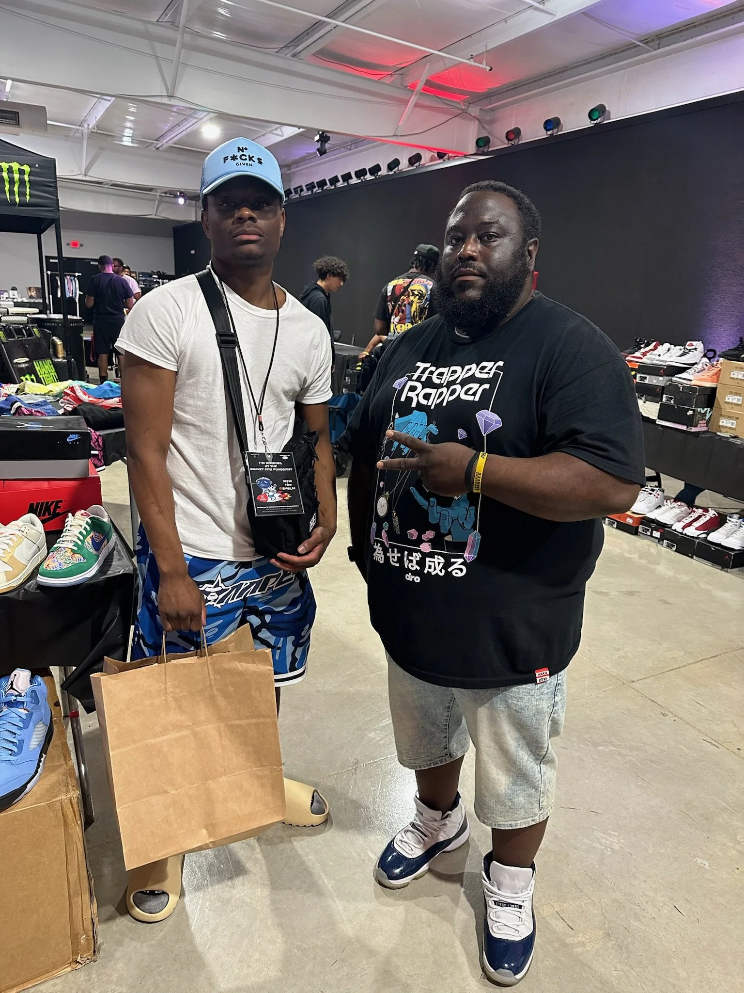 Two men standing in a sneaker store or event, with shoes and sneakers displayed on tables and shelves around them. One man is wearing a white t-shirt, blue patterned shorts, a blue cap, and holding a paper bag. The other man is wearing a black graphi