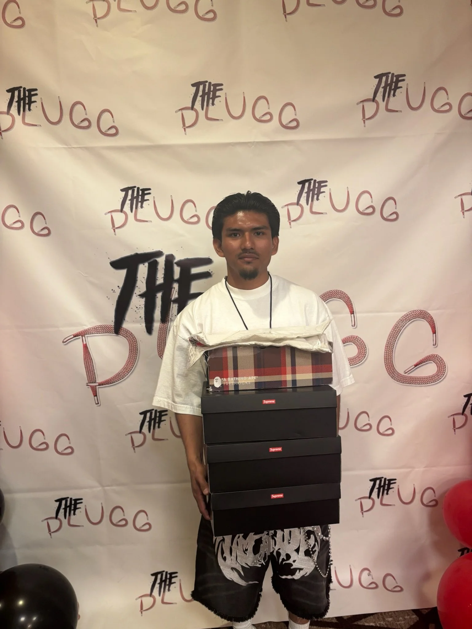 A young man standing in front of a backdrop with 'The Pugg' and 'The' written repeatedly. He is holding multiple shoeboxes stacked on top of each other, with a shirt draped over his shoulders.